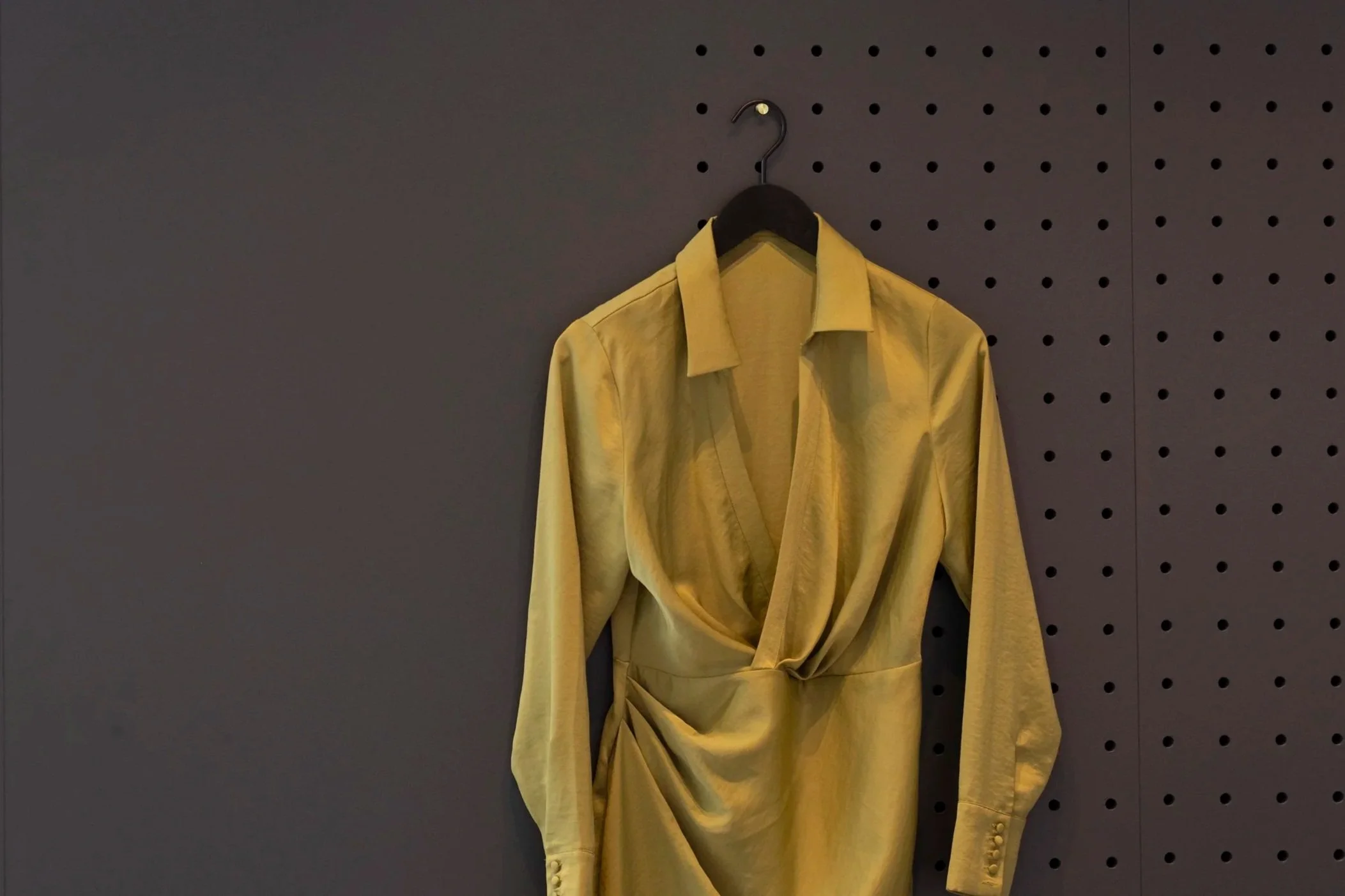 Yellow long-sleeve shirt or dress hanging on a black hanger against a gray pegboard wall.