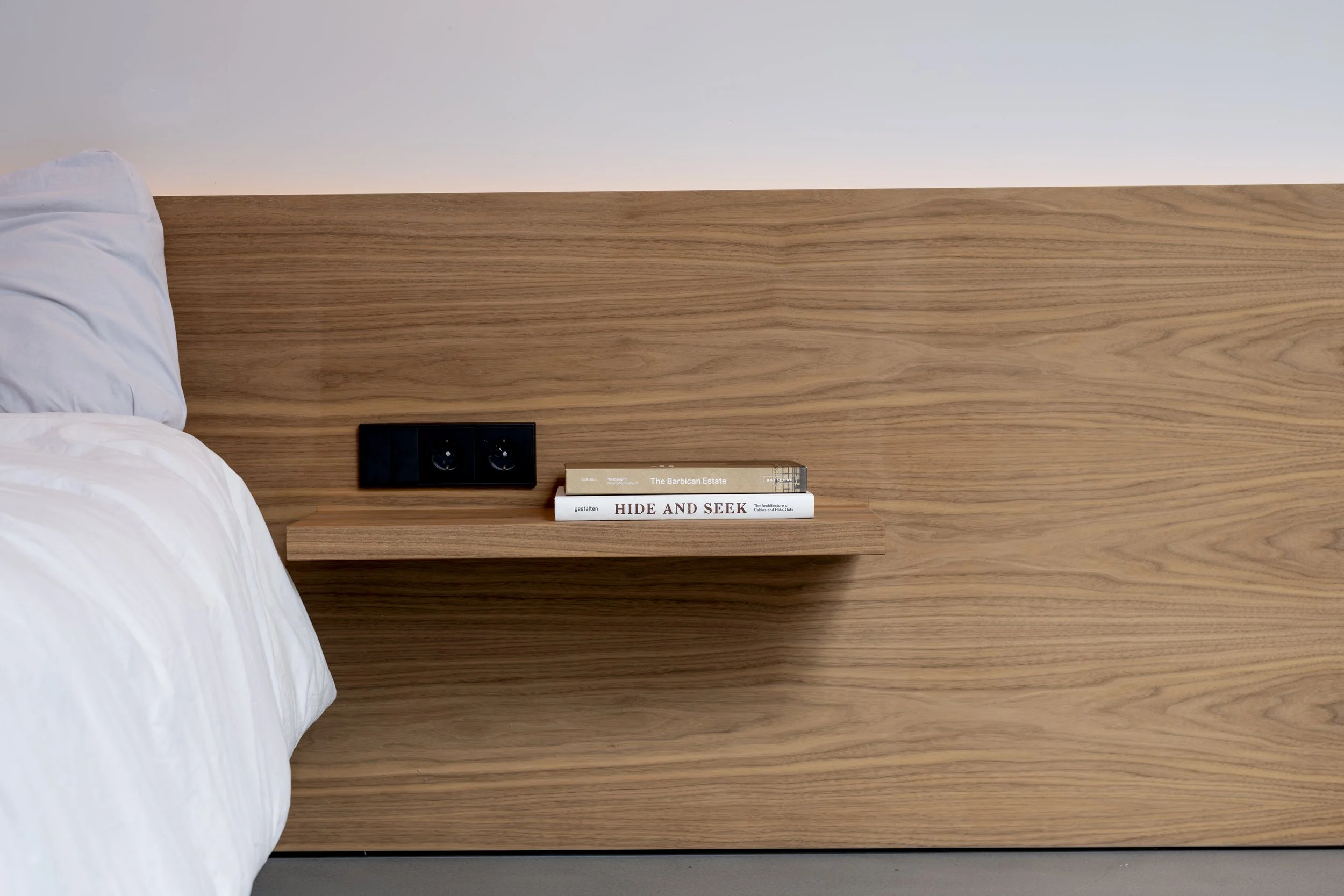 A close-up of a modern bedroom with a wooden headboard and a floating nightstand holding books, a black electrical outlet, and an off-white wall.