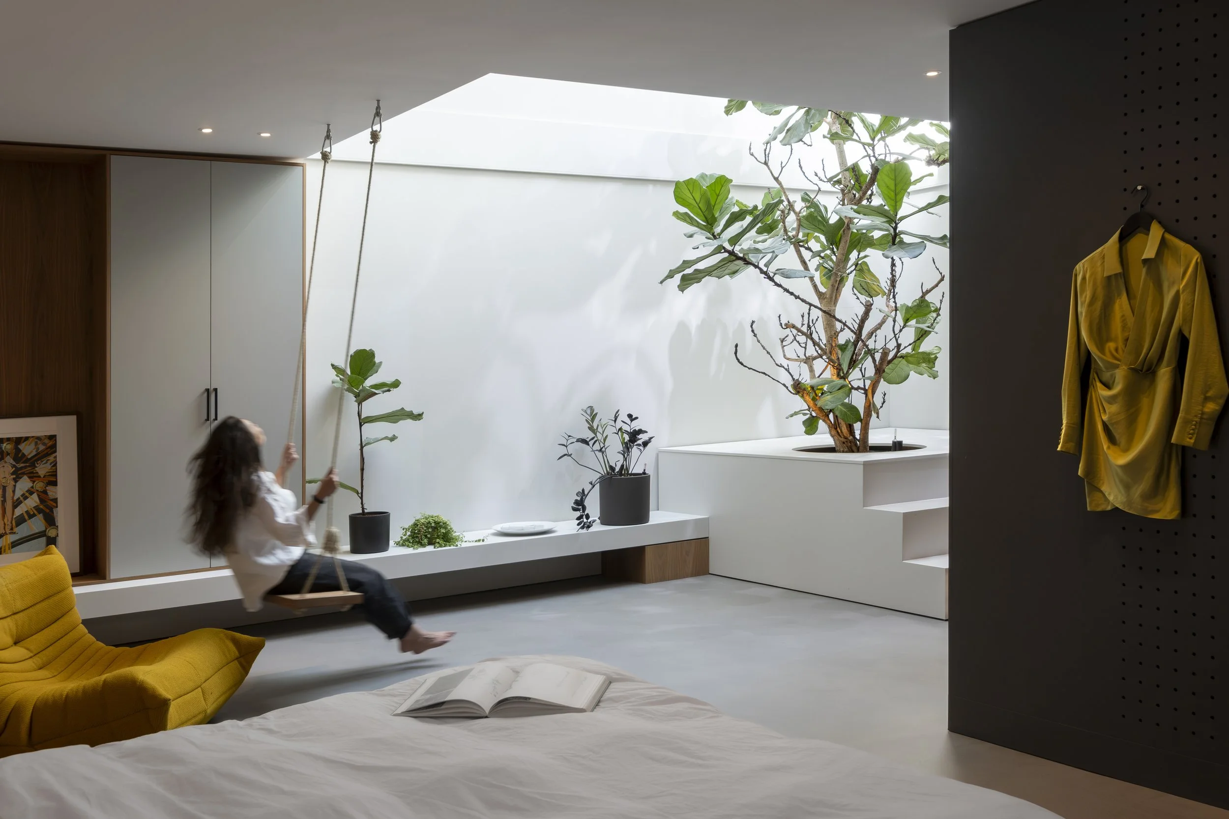Interior of a modern bedroom featuring a girl on a swing, a yellow chair, open book on a bed, potted plants, a large fig tree, and a black wall with a yellow shirt hanging, with natural light coming through a skylight.