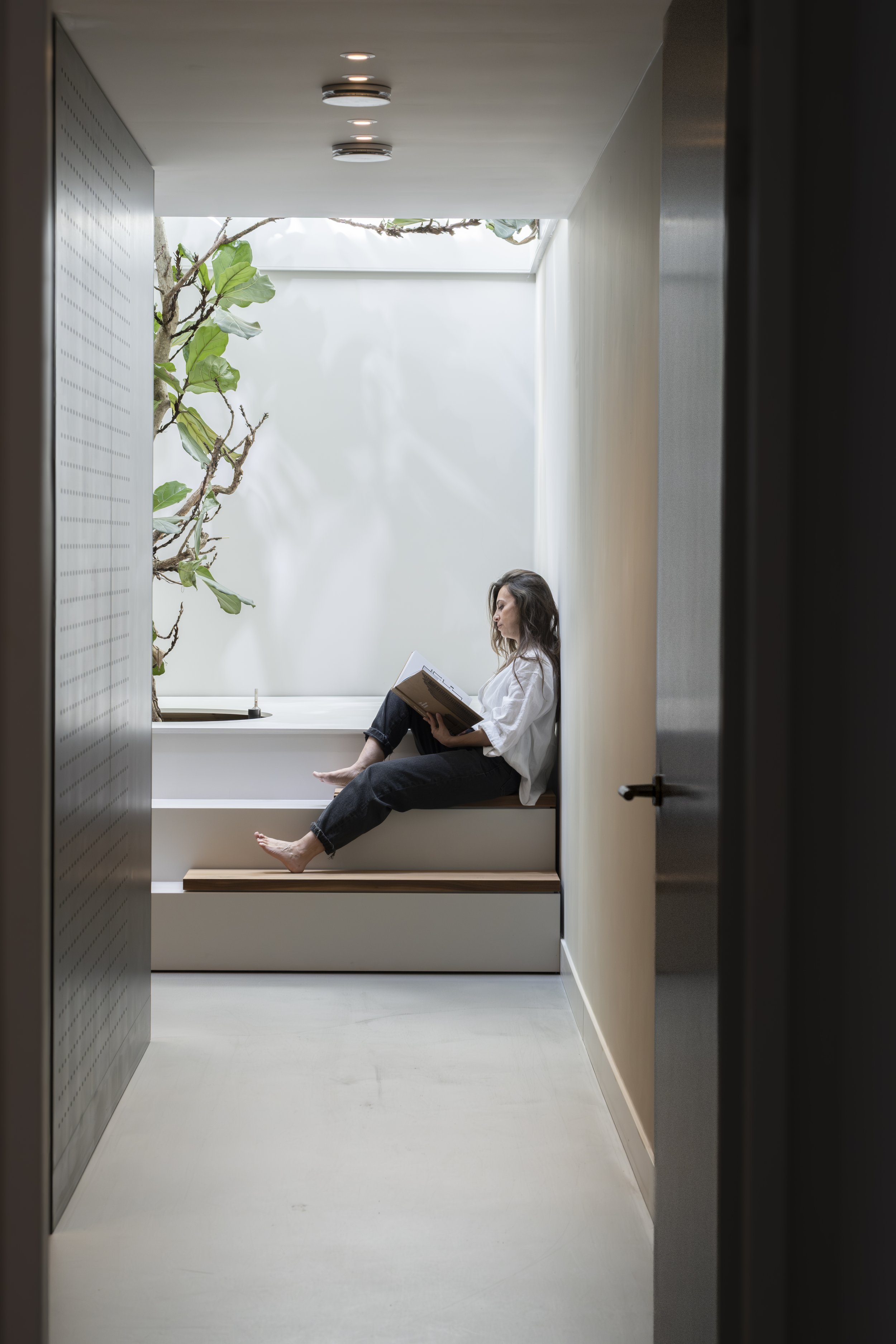 Woman sitting in a minimalist indoor space, reading a book by a small tree near a window with natural light.