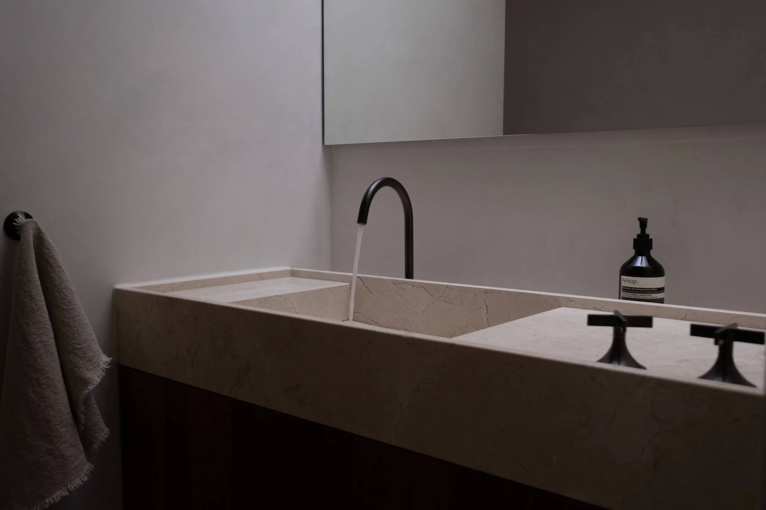Minimalist bathroom with beige marble countertop sink, black faucet, mirror, gray towel hanging on wall, and black bottle of Aesop hand soap.