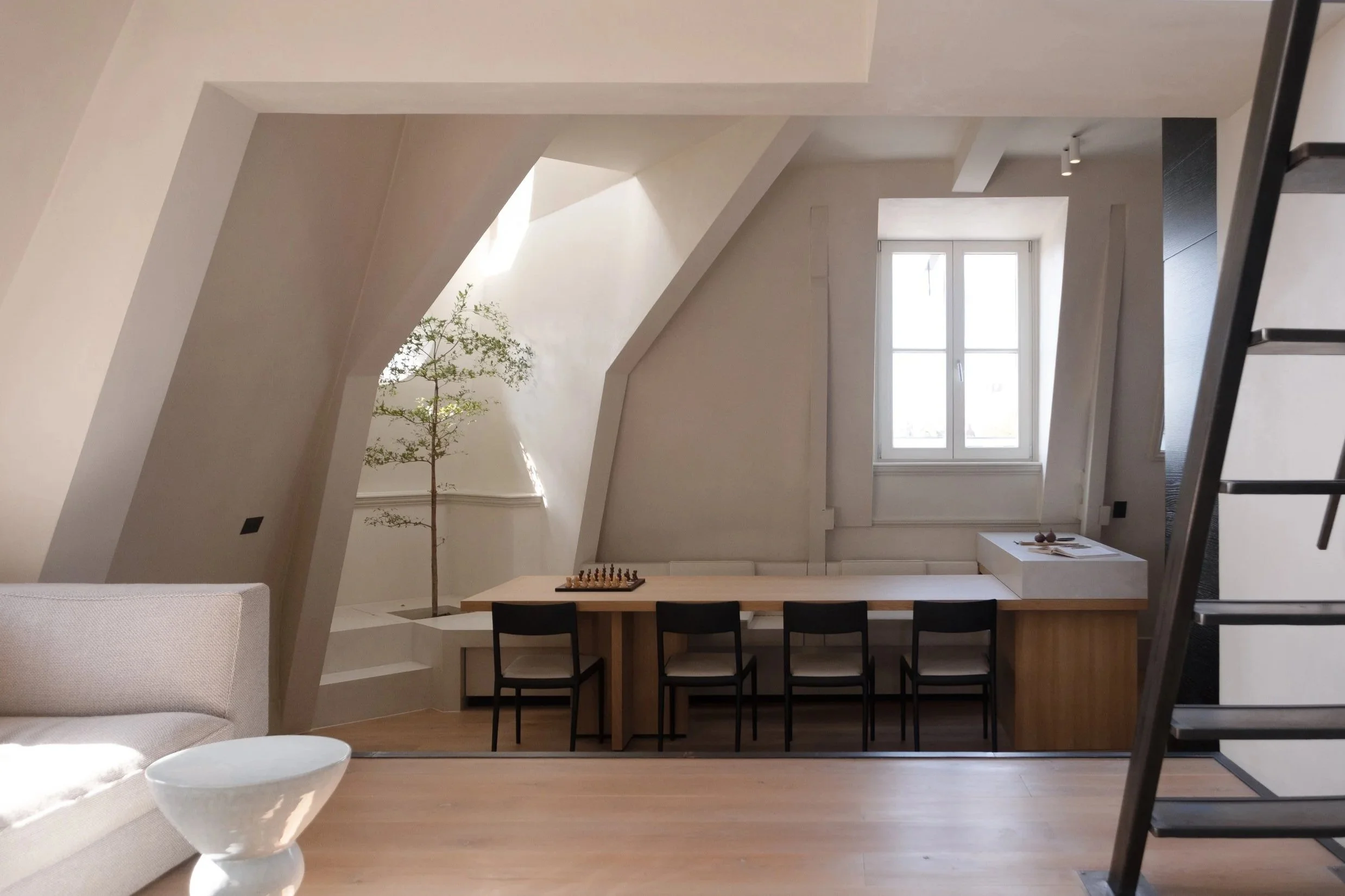Minimalist living room with a beige sofa, a white oval coffee table, and a long wooden table with four black chairs near a window. A small tree is on a ledge next to the wall.