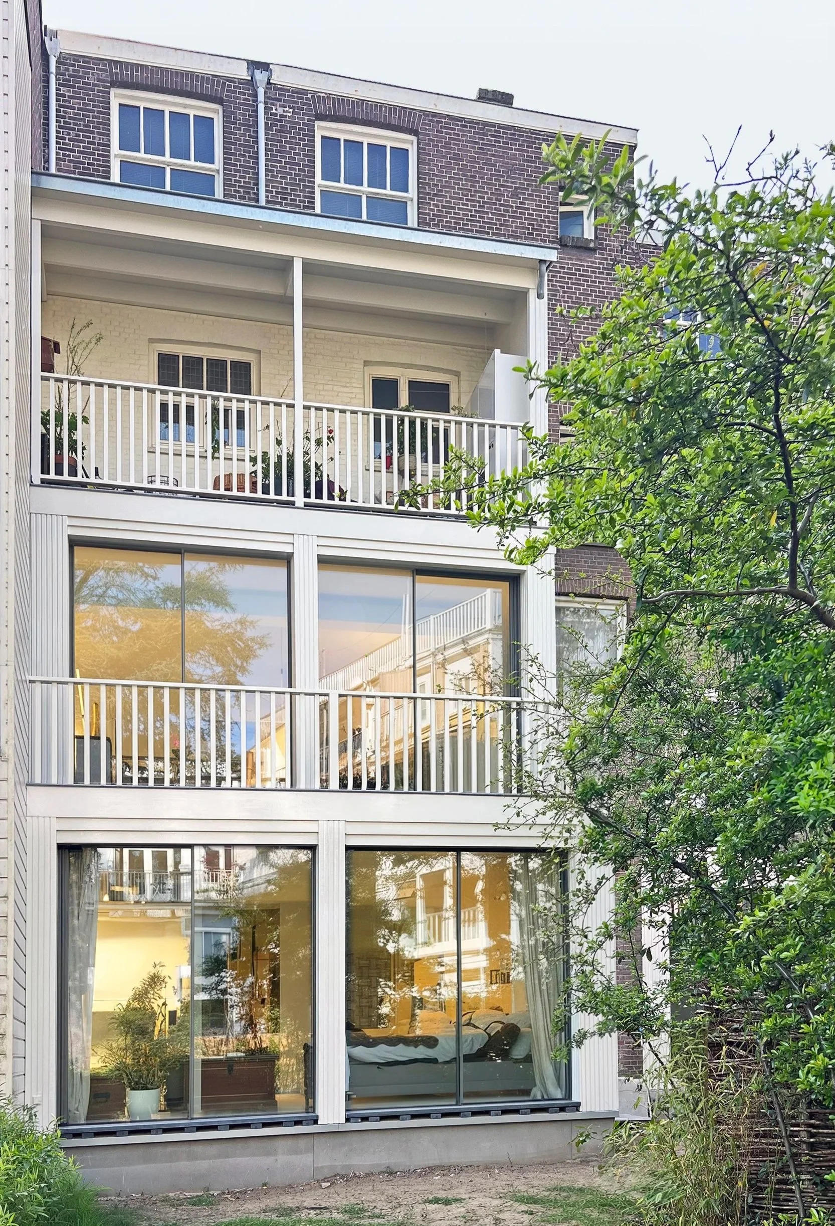 Multi-story residential building with large glass sliding doors on the ground floor, a balcony with plants on the second floor, and brick exteriors. Trees and greenery are in the foreground.