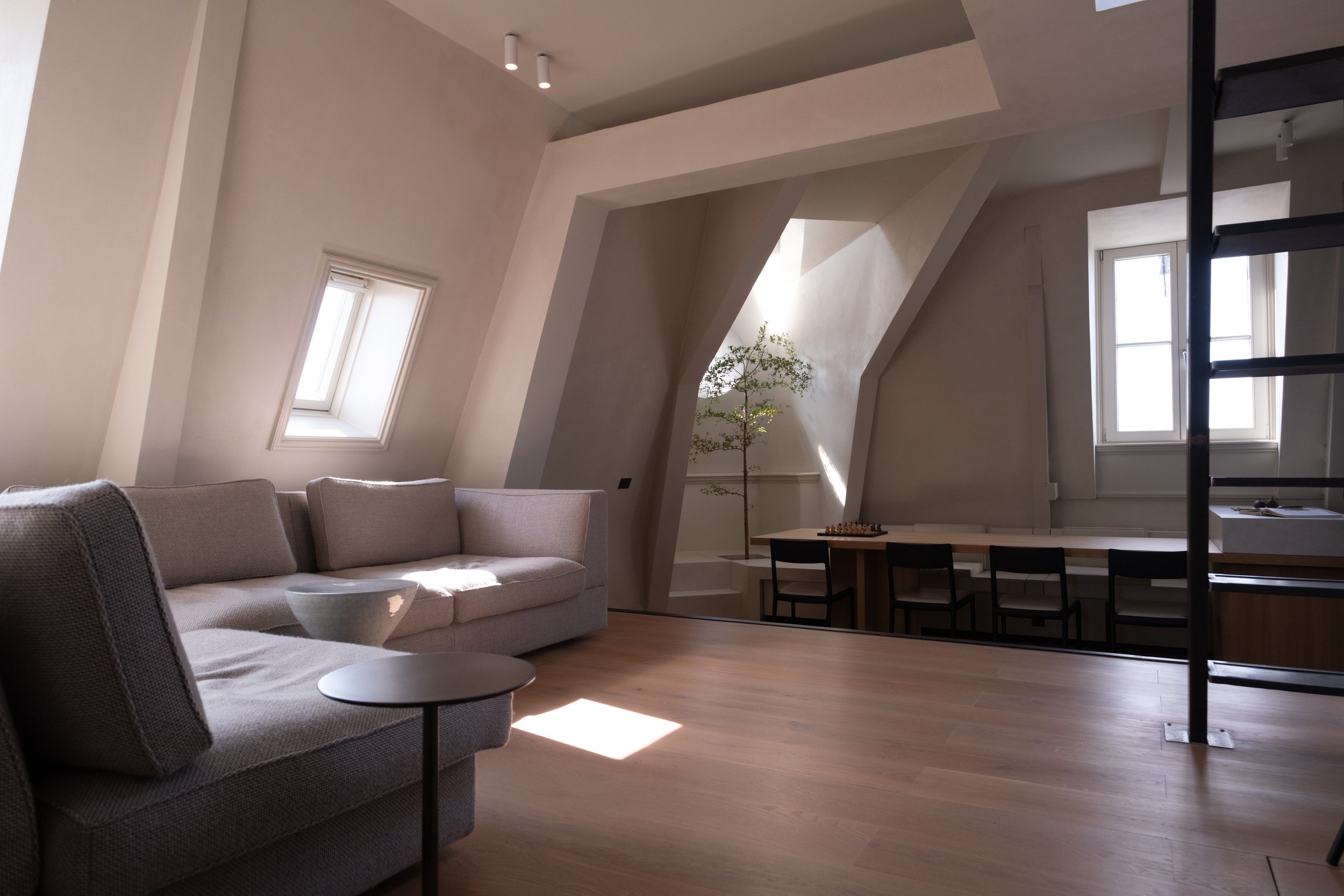 Modern living room with angled ceilings, two windows allowing natural light, a beige couch, small side tables, a potted tree, and a wooden ladder.