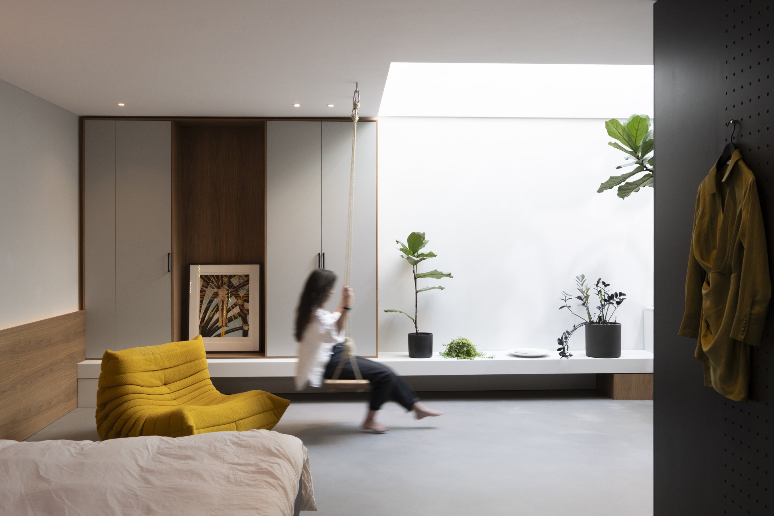 A modern room with a girl on a swing, yellow armchair, potted plants on a ledge, framed artwork, bed in foreground, and a yellow jacket hanging on a black pegboard.