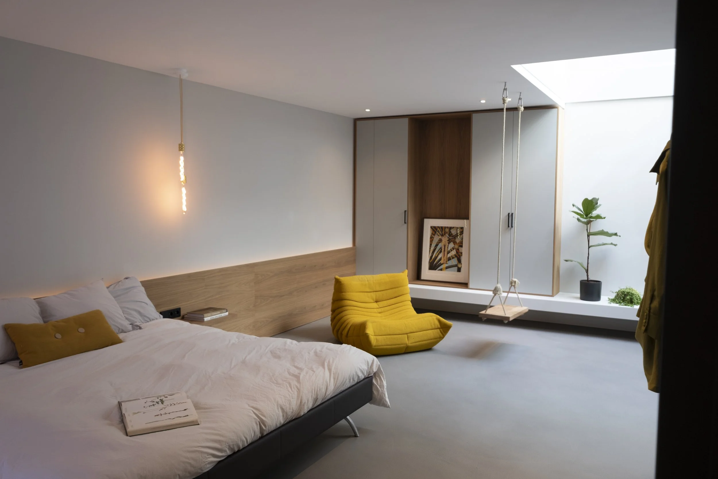 Modern bedroom with a bed, yellow accent pillow, yellow lounge chair, framed artwork, hanging swing, and potted plants near a skylight window.