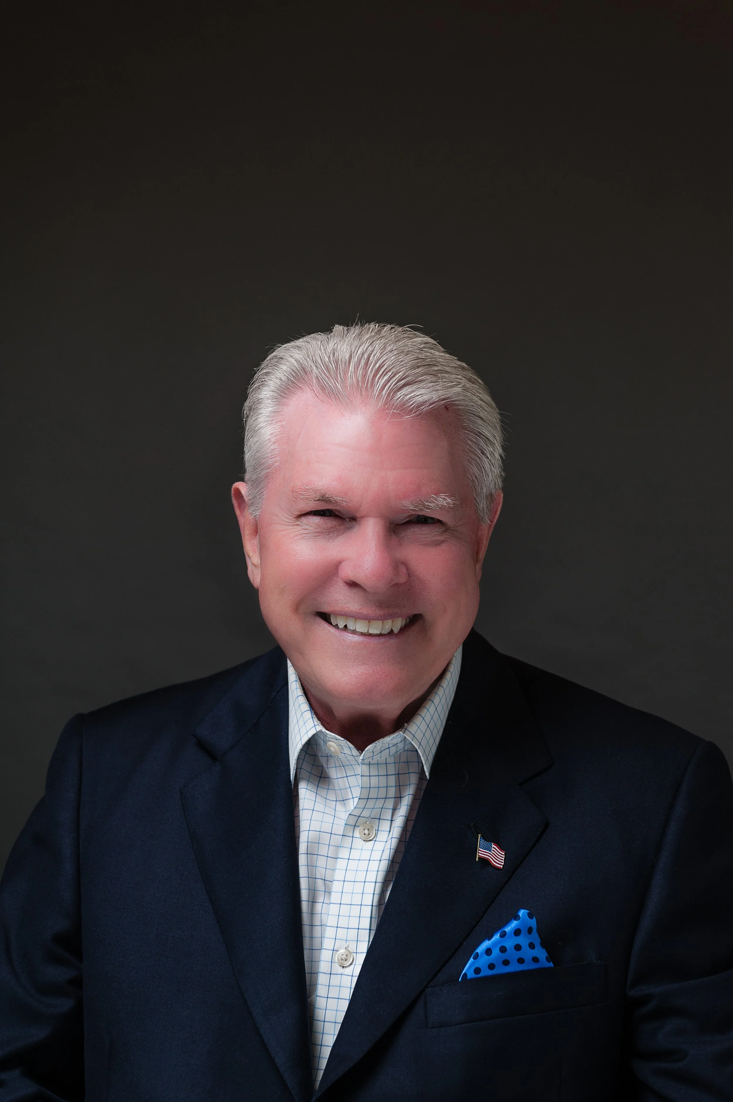 A smiling man with gray hair wearing a dark suit, white checkered shirt, and blue polka dot pocket square, against a plain dark background.