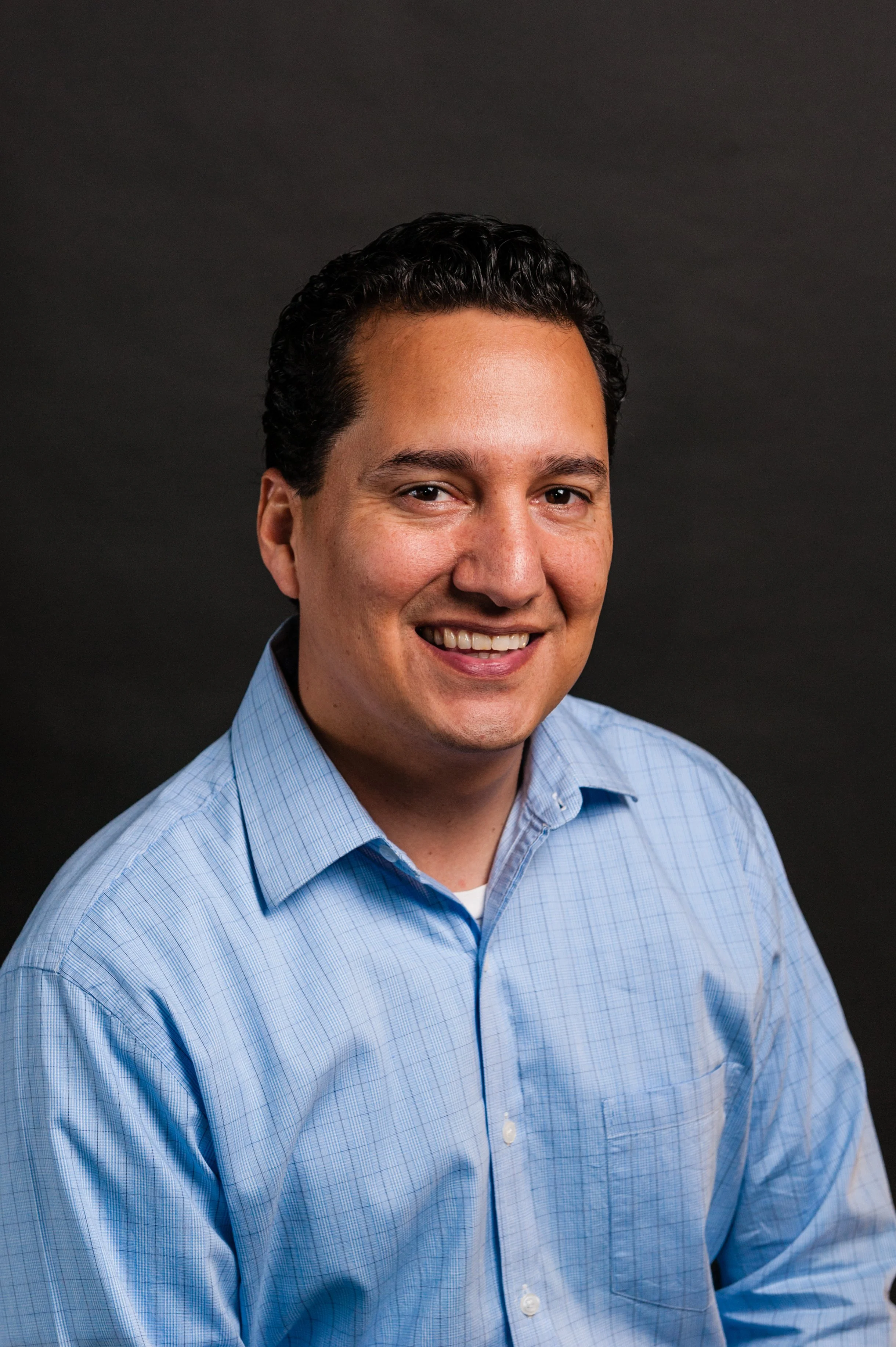 A portrait of a smiling man in a blue checked shirt against a dark background.