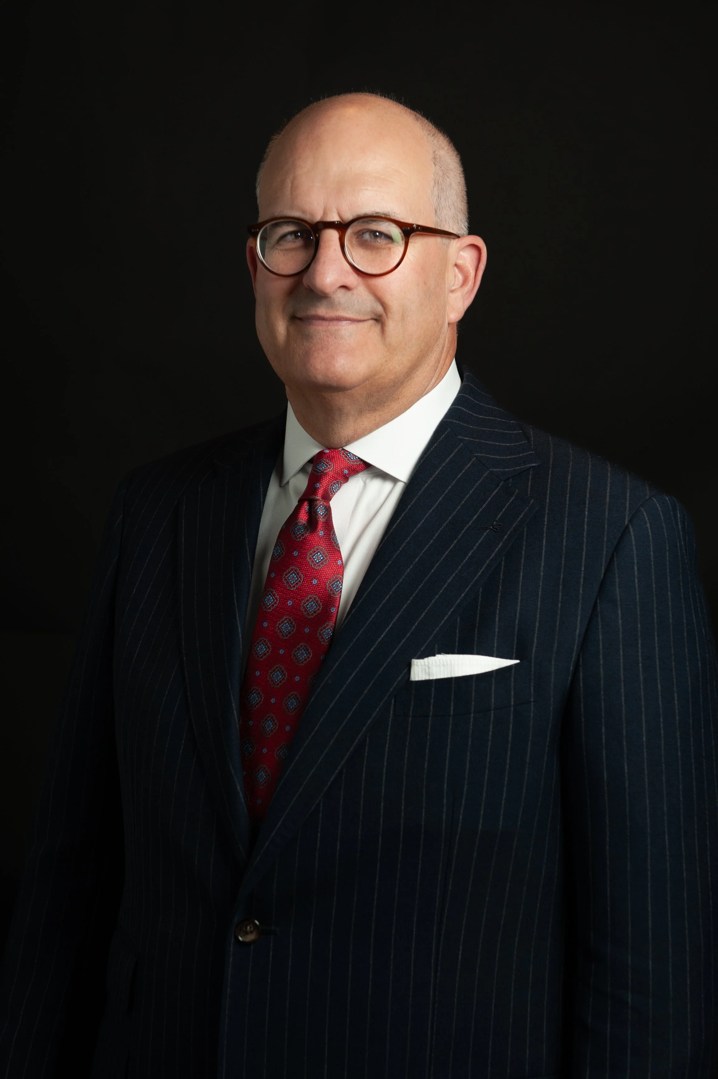 A middle-aged man wearing glasses, a dark striped suit, a white shirt, and a red patterned tie, standing against a black background.