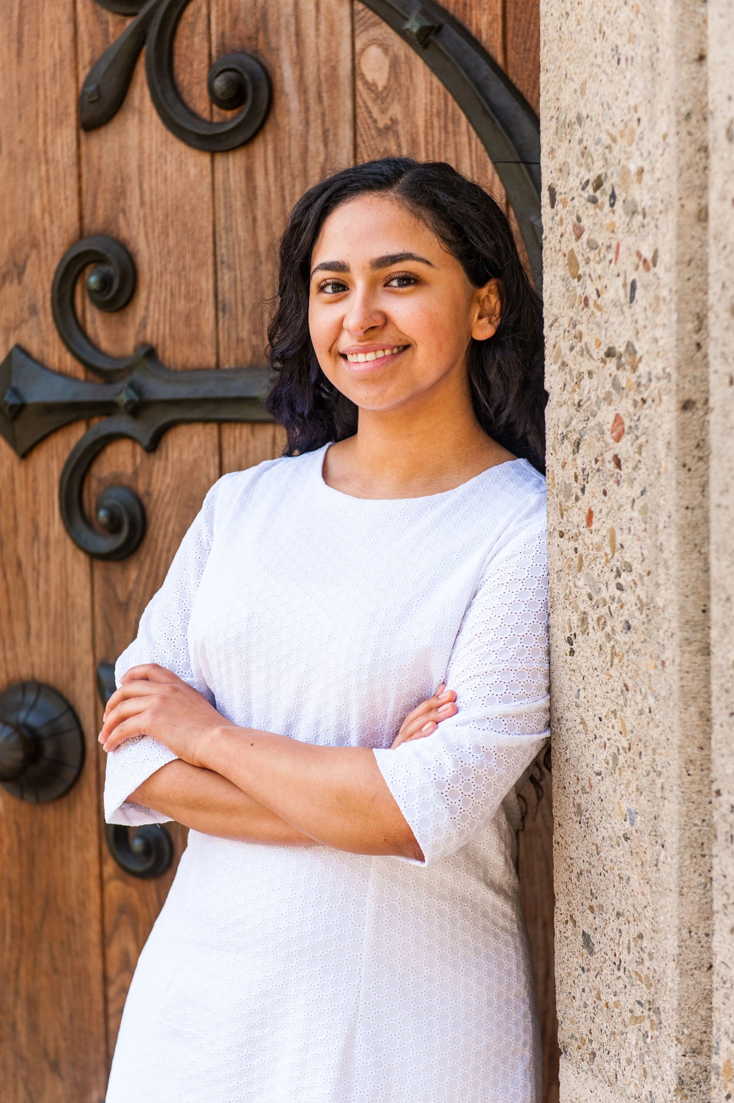 A young woman with dark curly hair and a smile, wearing a white dress, standing beside a stone wall and wooden door with decorative black wrought iron accents.