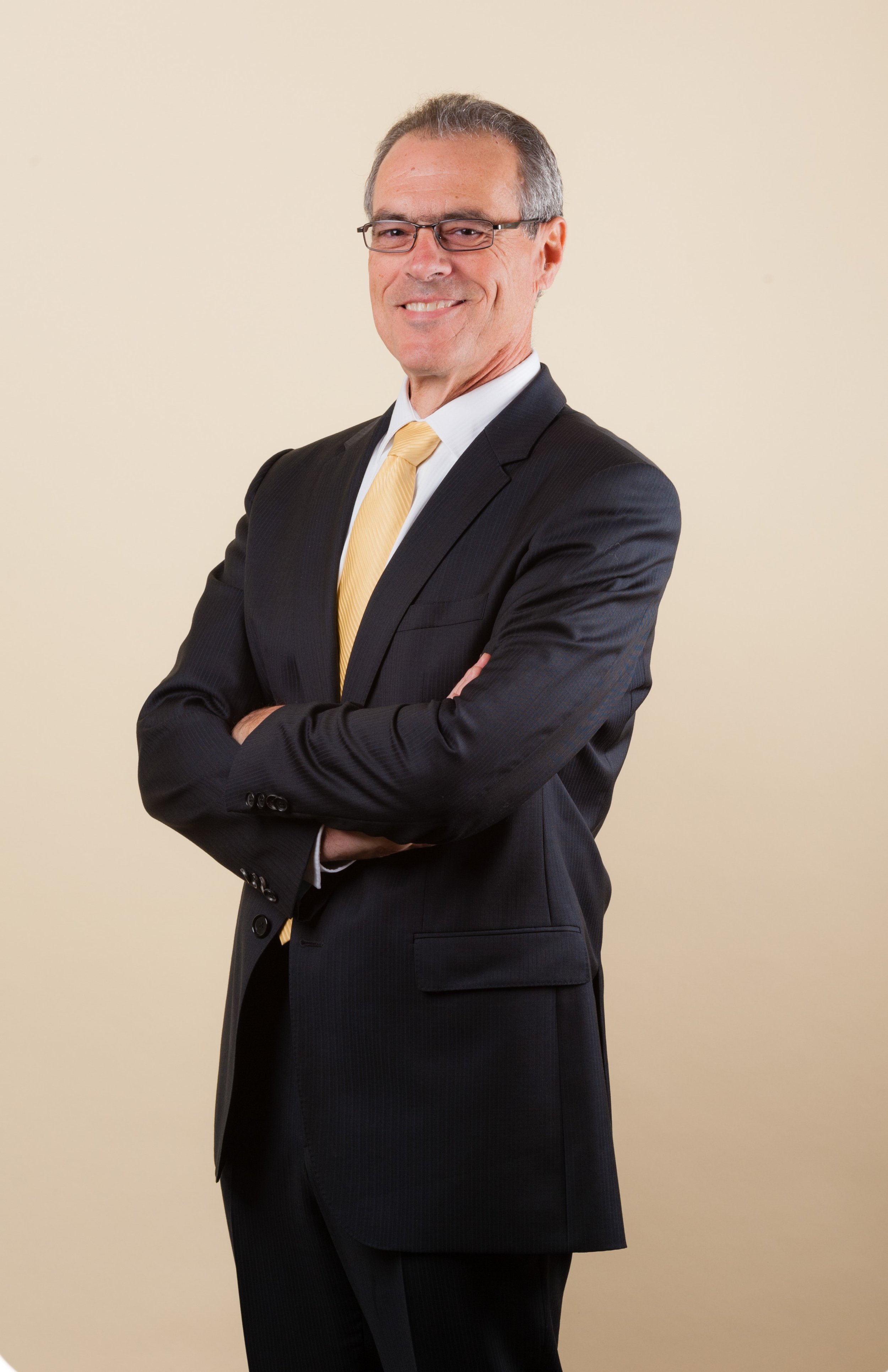 A middle-aged man in a black suit with a yellow tie, wearing glasses, smiling with his arms crossed against a beige background.