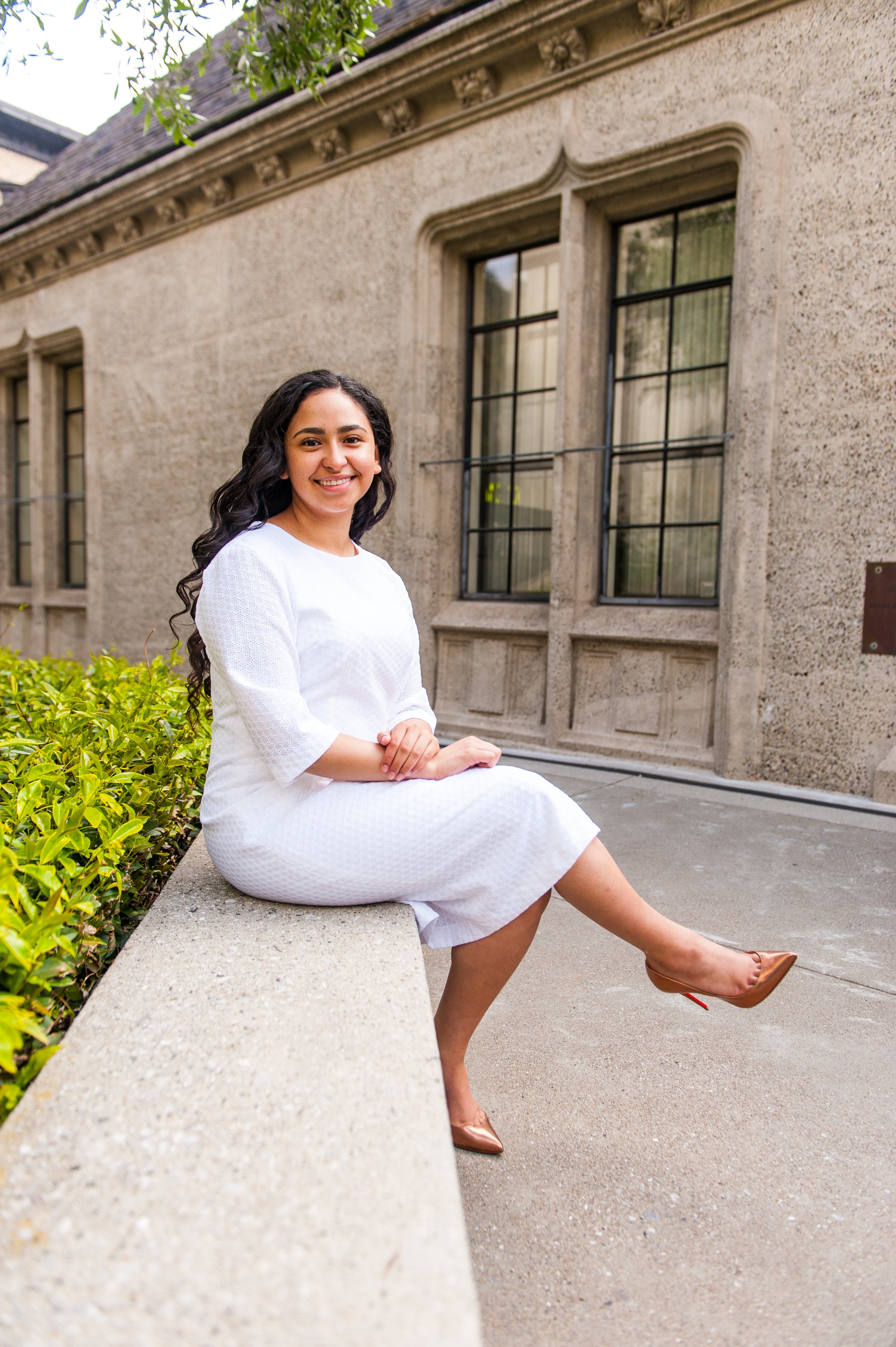 Young woman with long, dark curly hair sitting on a concrete ledge outdoors in front of a historic building with beige stone walls and large windows, smiling at the camera.