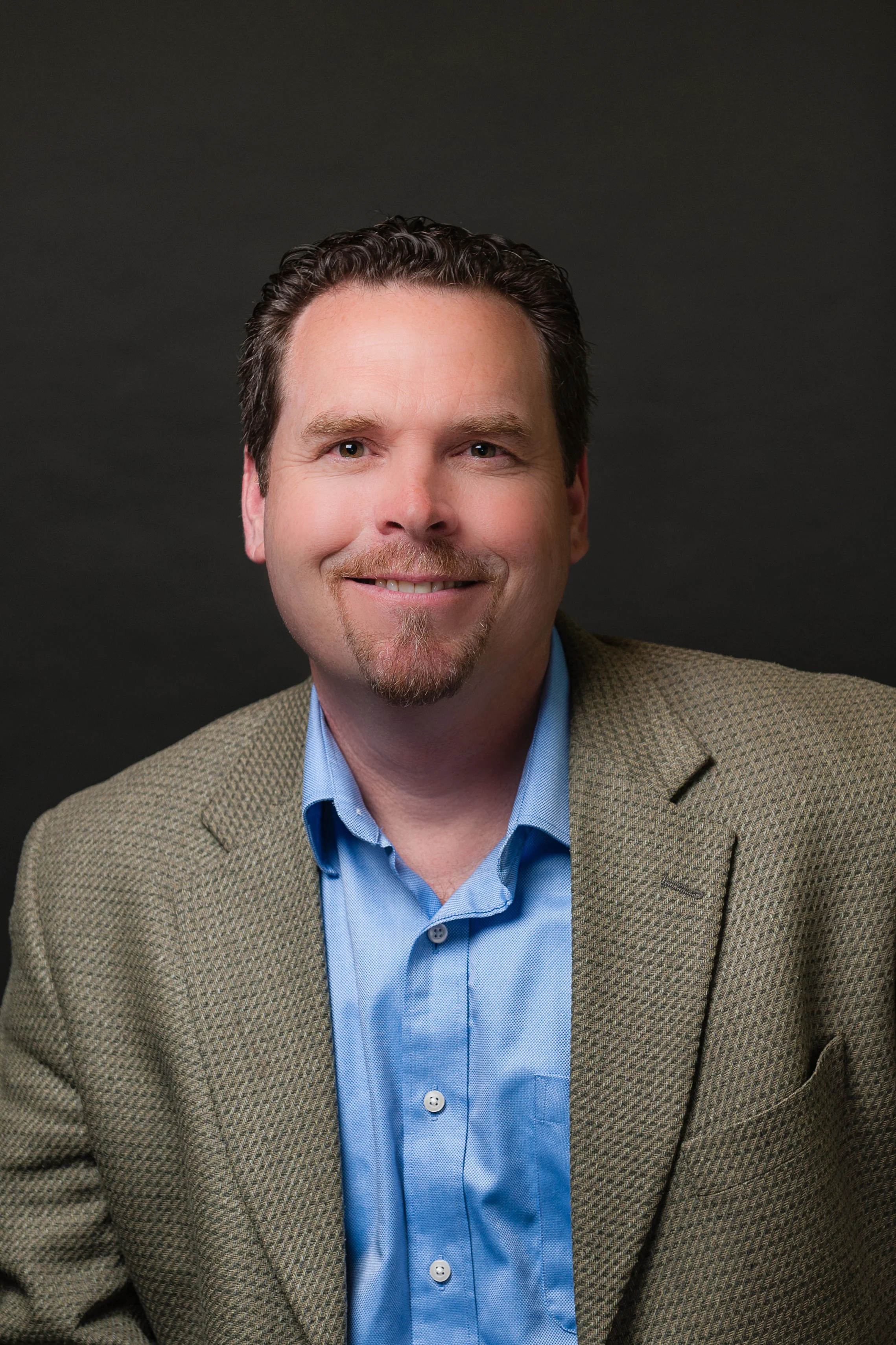 Portrait of a man with dark hair and a goatee, smiling, wearing a beige blazer and a blue dress shirt against a dark background.