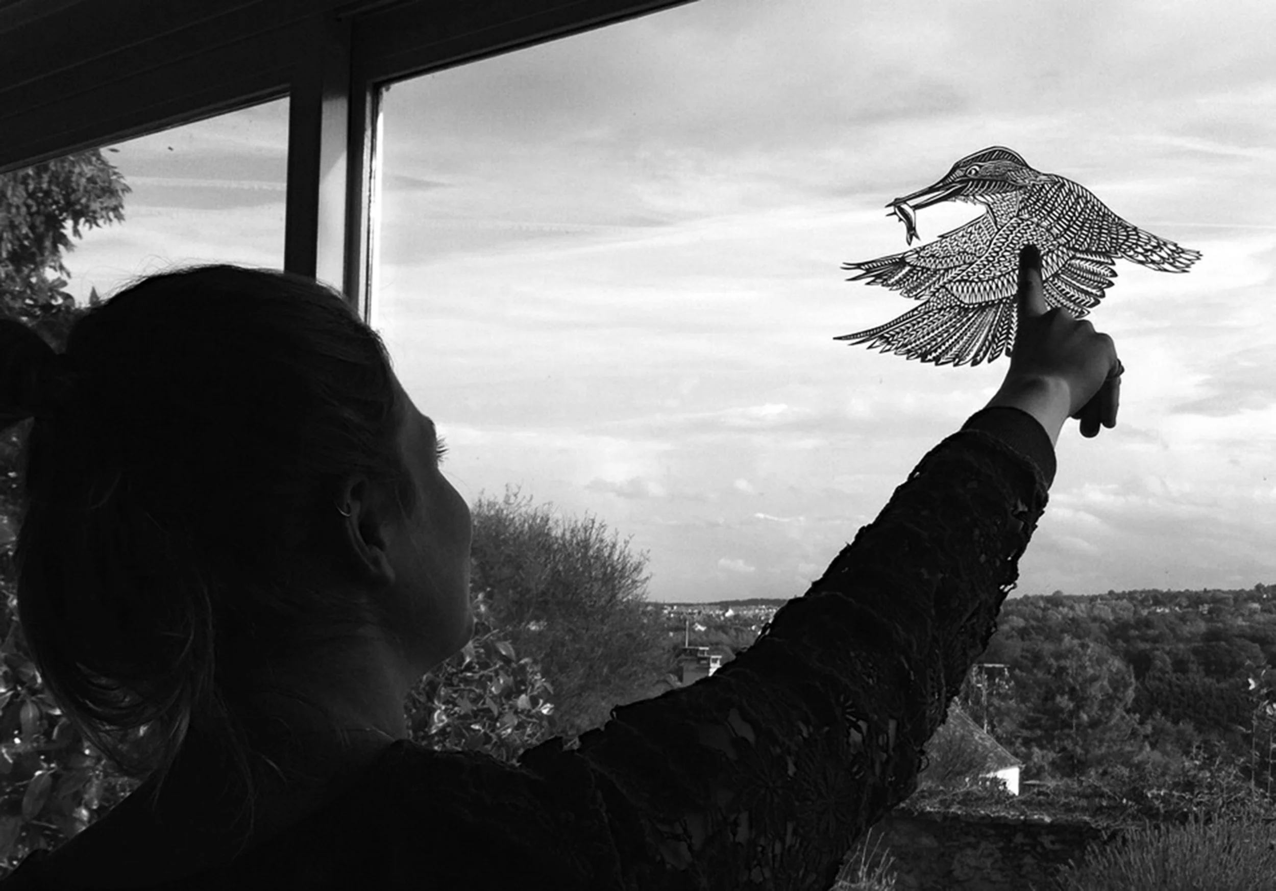 The artist herself holding a kingfisher papercutting infront of a window
