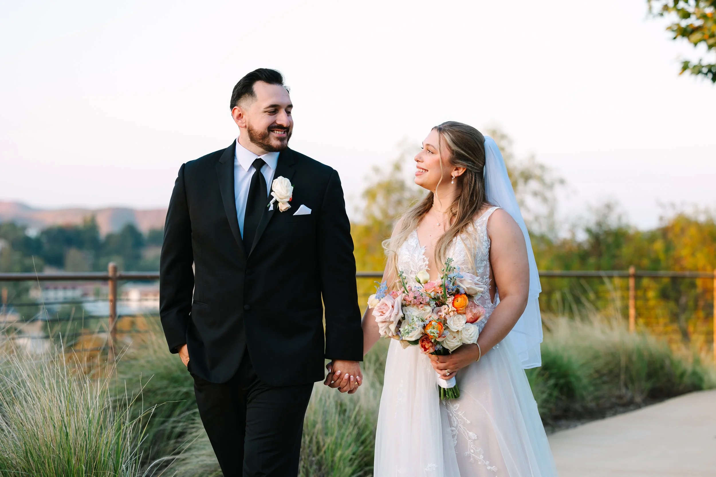 A bride and groom smiling outdoors in a garden during sunset, with the bride holding a colorful bouquet of flowers and the groom wearing a black suit with a boutonniere. Agoura Hills Recreation and Event Center Wedding CA