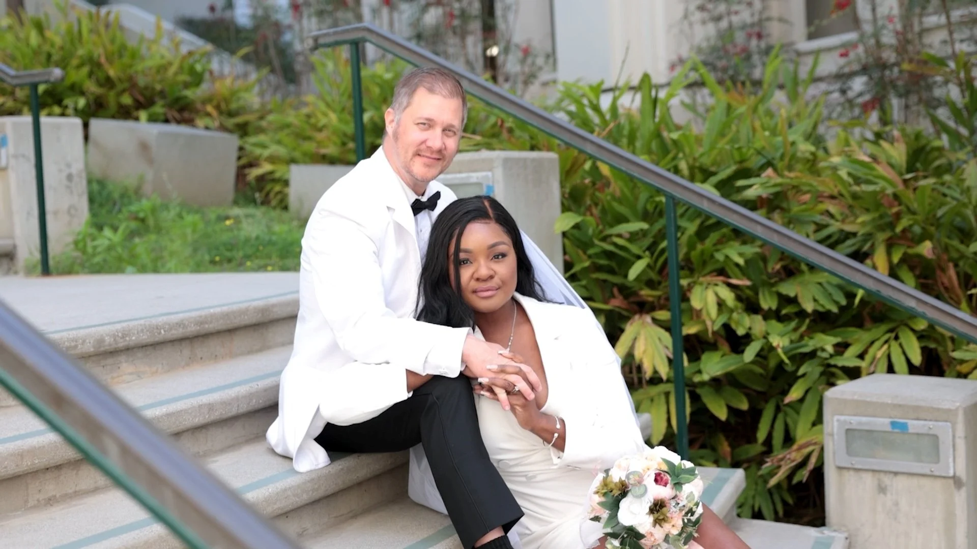A couple in wedding attire sitting on outdoor stairs, surrounded by greenery, with the groom in a tuxedo and the bride in a white dress holding a bouquet.