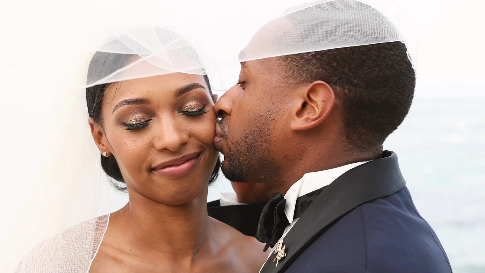 A bride and groom sharing a kiss, with the groom gently kissing the bride's cheek, both wearing wedding attire and with a blurred sky background.
