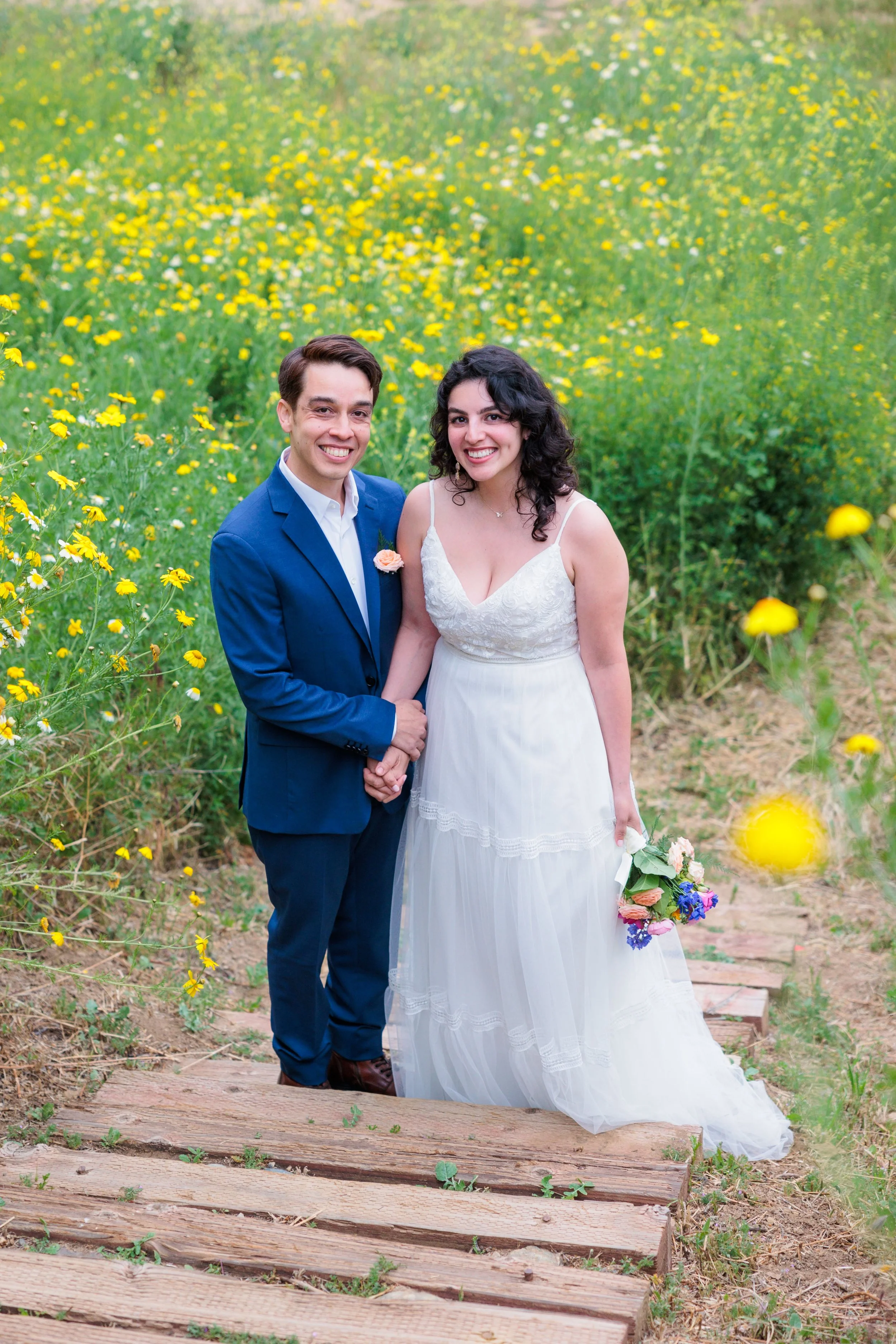A couple in wedding attire standing on a wooden path in a field of yellow flowers, smiling at the camera.