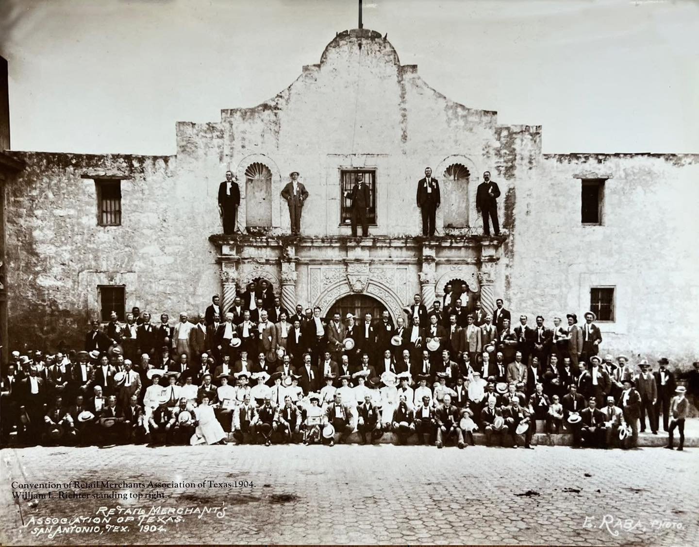 Take a moment to really look at this photo.
It shows the Retail Merchants Association of Texas convention gathered at the Alamo in 1904. One detail stands out right away. People are standing directly on the building itself, something that would never