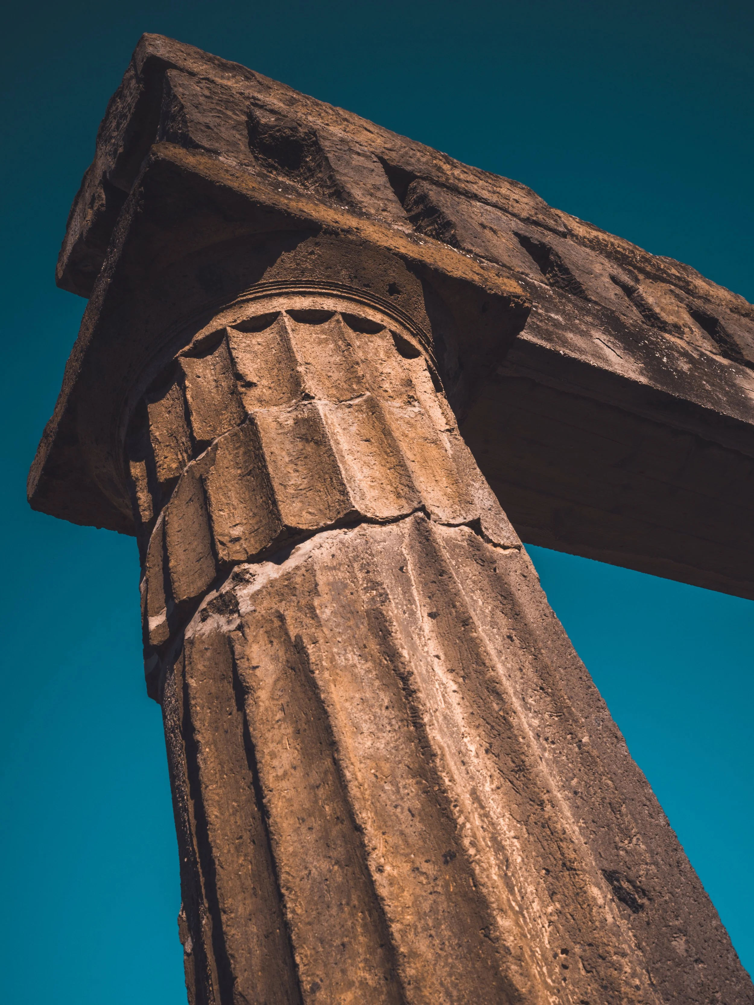 Close-up of an ancient stone column with a decorated capital, part of a historical building or temple, against a clear blue sky.