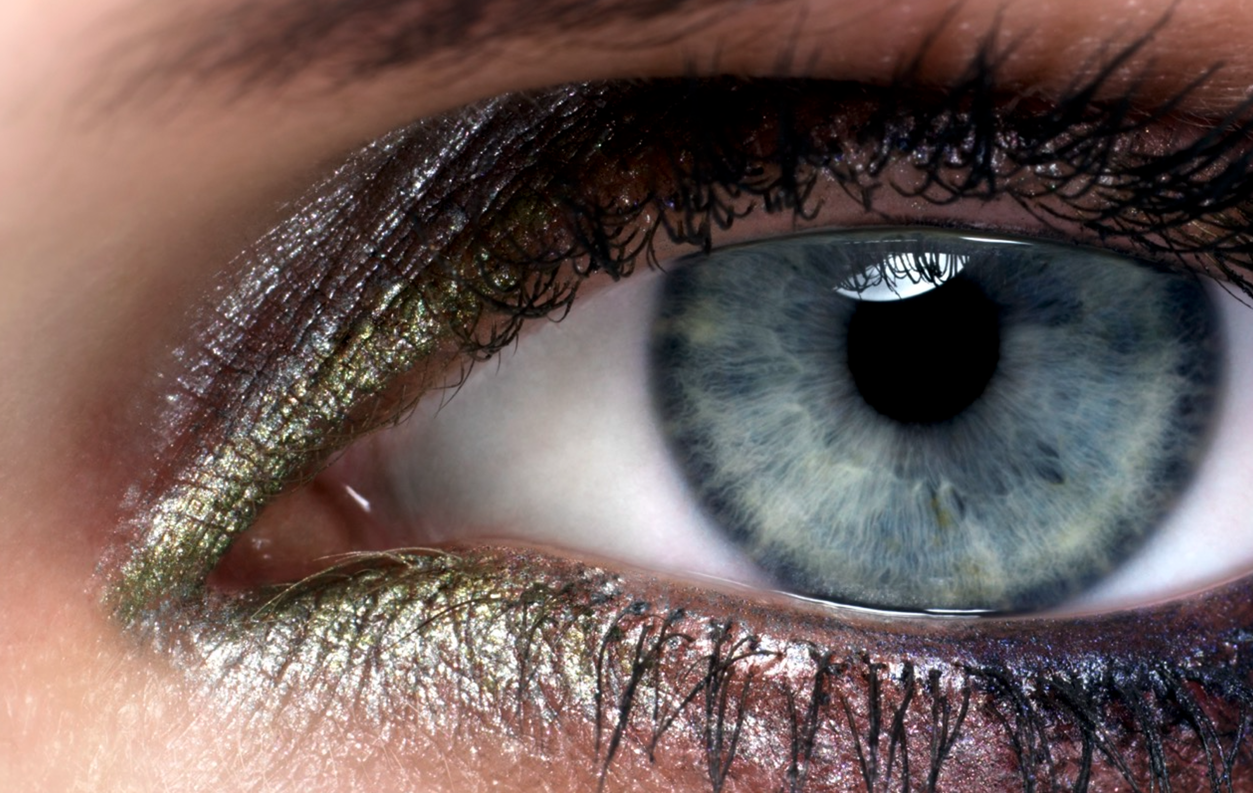 Close-up of a human eye with grayish-blue iris, accented with gold eyeshadow, eyeliner, and mascara, surrounded by detailed eyelashes and skin with slight shimmer.
