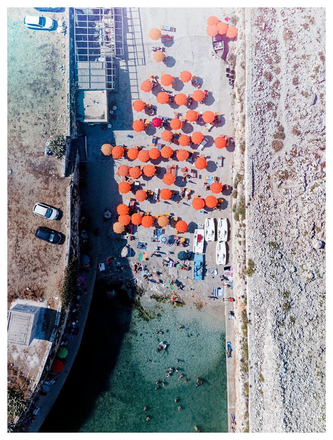 Bird's-eye view of a beach with orange umbrellas, lounge chairs, and people swimming in a small body of water, adjacent to a rocky shoreline and parking lot with cars.