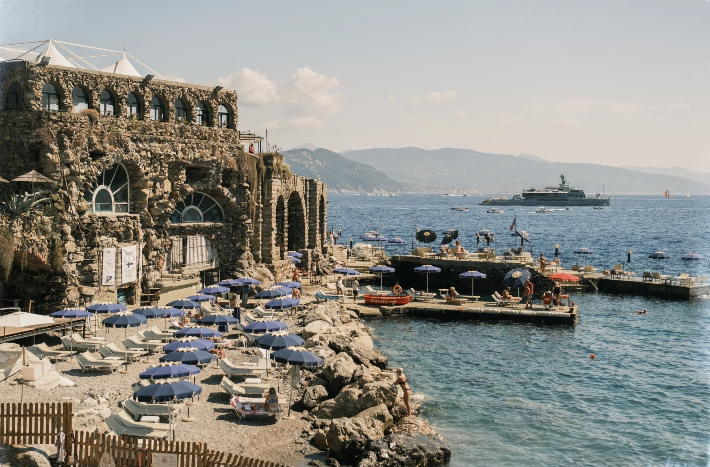 A scenic coastal view featuring a rocky beach with numerous blue umbrellas and lounge chairs, a large yacht in the water, and historic stone buildings perched on the shoreline, with hills and mountains in the background on a clear day.