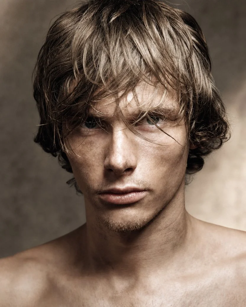 A close-up portrait of a young man with messy, shoulder-length brown hair and light skin. He has a serious expression, with slightly pursed lips and intense eyes, against a plain background.