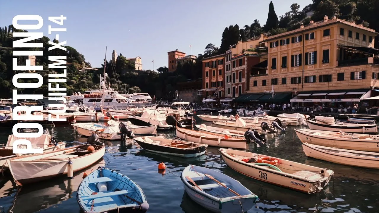 A harbor filled with various small boats docked near a waterfront restaurant, with colorful buildings and hillside trees in the background, during daylight.