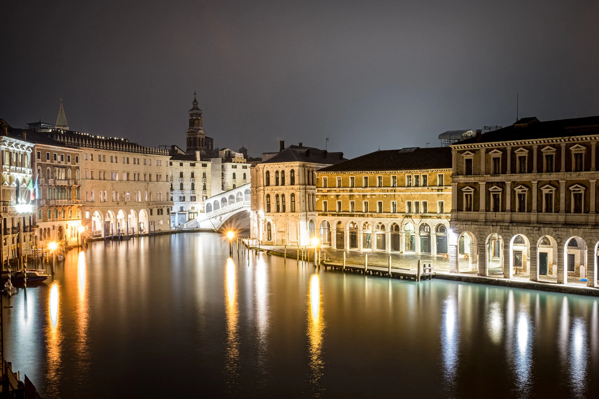 Night view of historic buildings along a canal in Venice, Italy, with calm water reflecting the illuminated architecture and dark sky overhead.