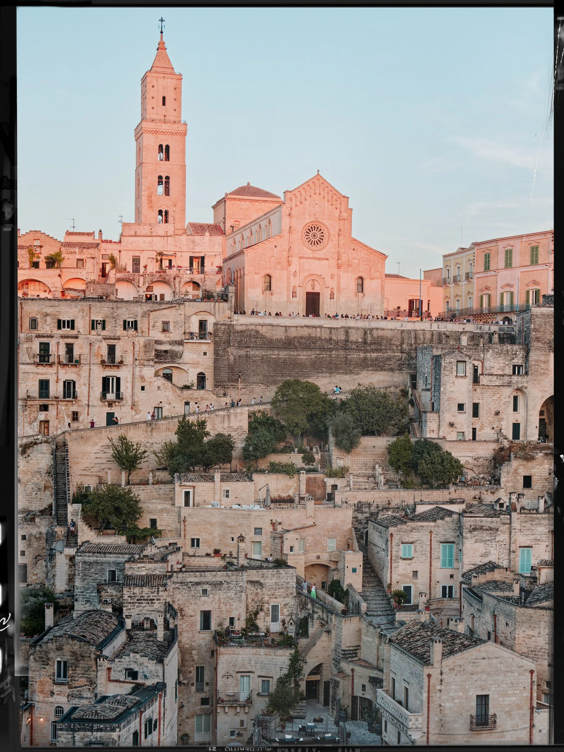 A historic European cityscape with a large stone church featuring a rose window and a bell tower, surrounded by ancient stone buildings on various levels, with some trees and a small outdoor seating area visible at the bottom.