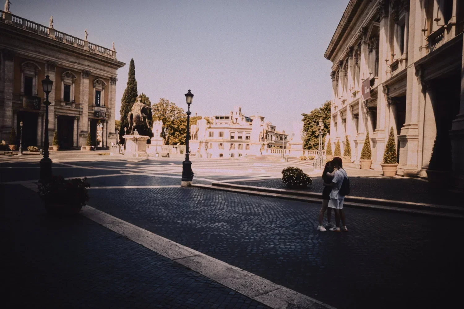 A couple stands close together on a cobblestone street in front of grand historic buildings with columns and statues, with trees and lampposts in the background.