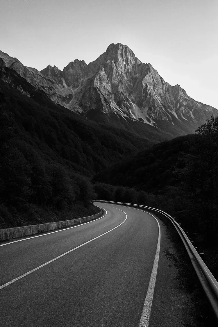 A winding road through a mountainous area with a large mountain peak in the background, captured in black and white.