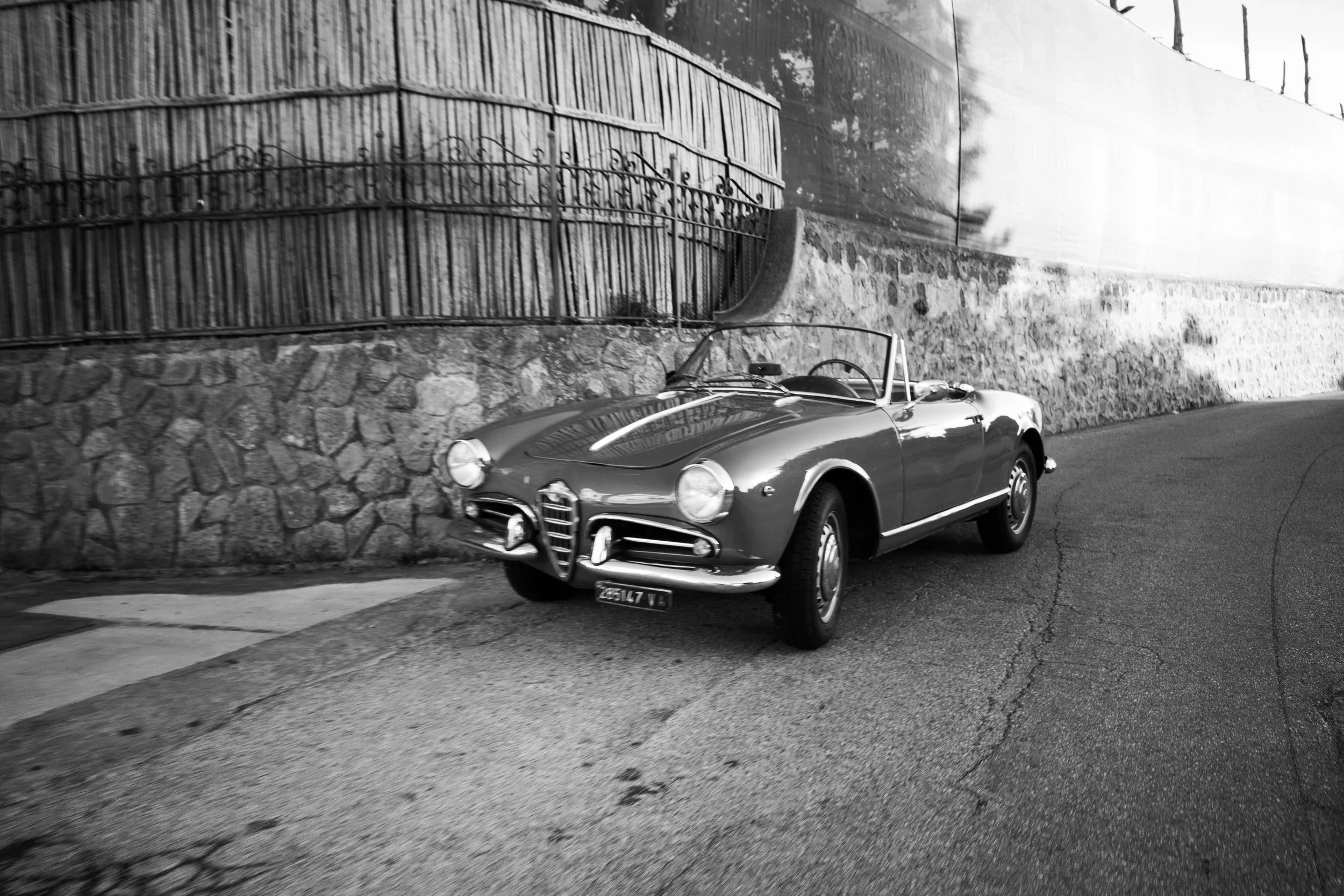 A vintage convertible car parked next to a stone wall with a metal fence on top, along a street with a sidewalk.