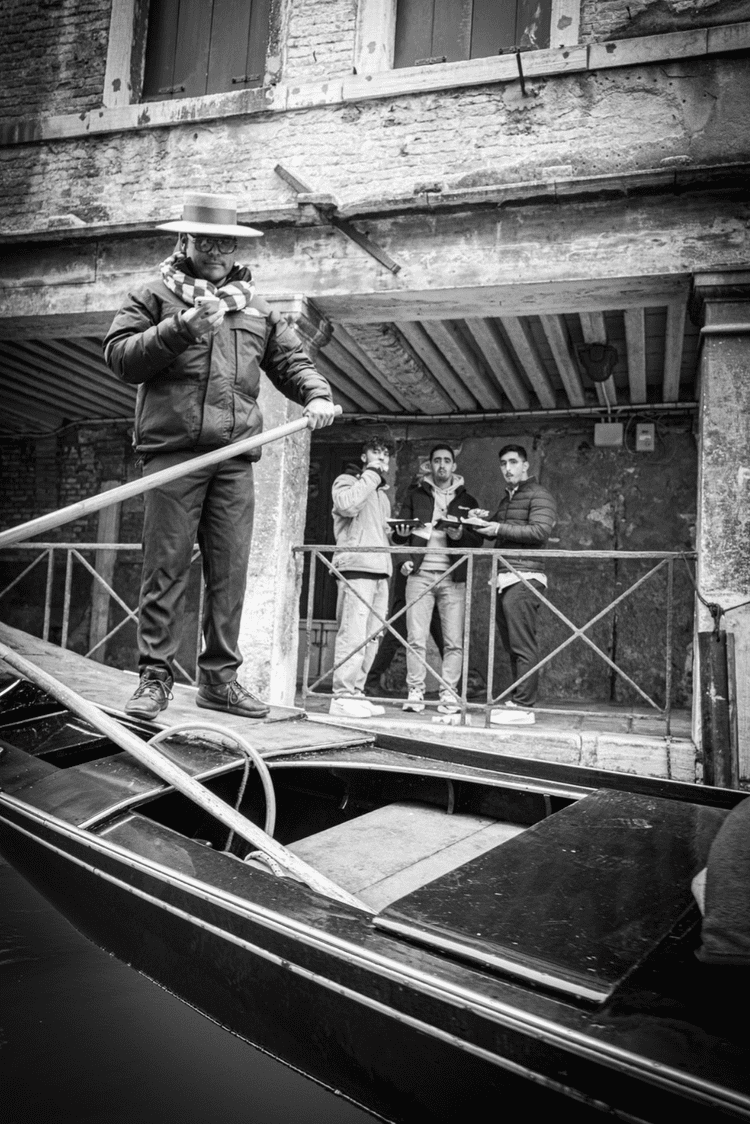 Four men on a dock near a boat, with one man standing on the boat holding a pole and using a smartphone, while the other three are on the dock eating food and looking at the camera, with an old brick building in the background.