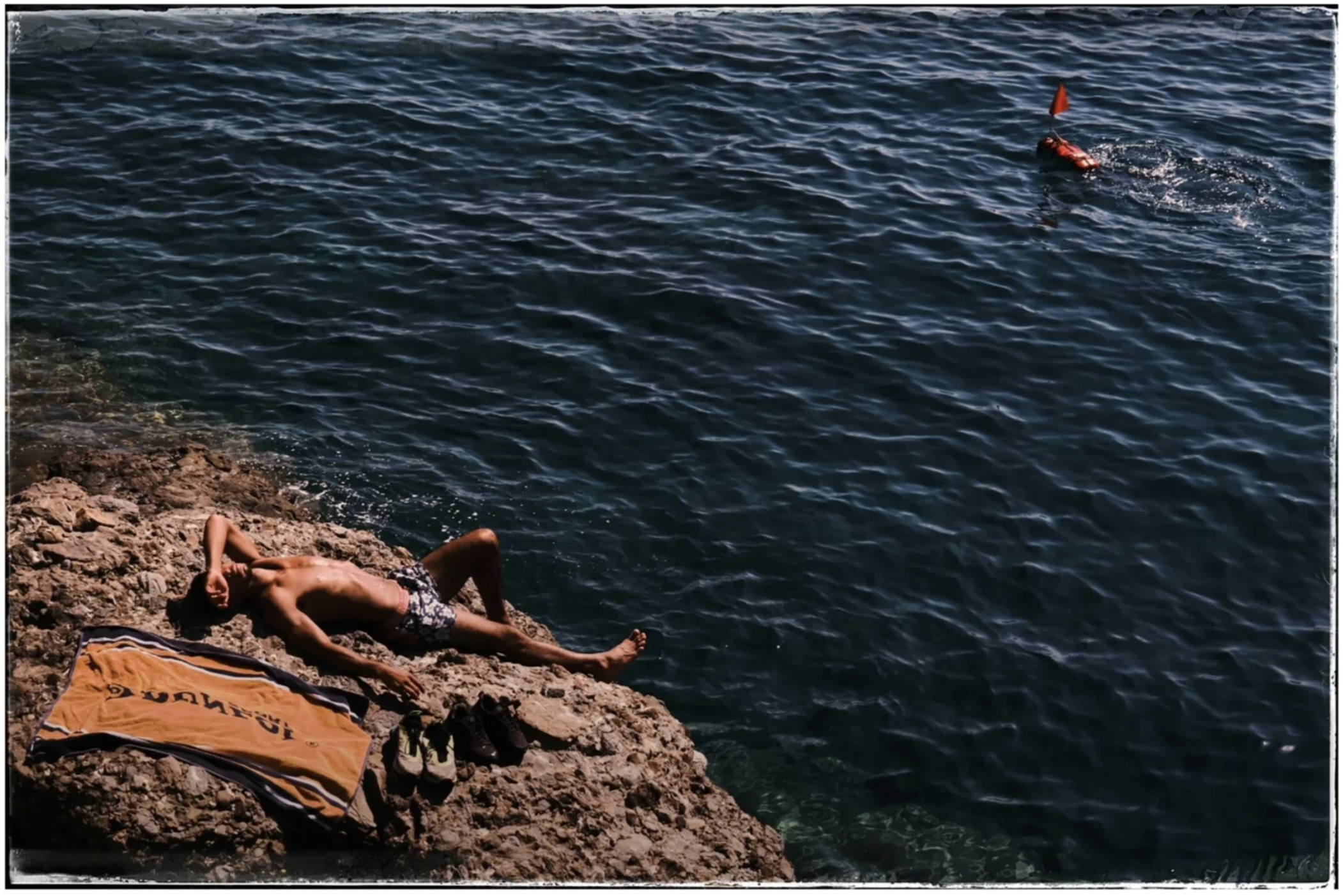 A person lying on rocks near the ocean, sunbathing on a towel with shoes nearby, with water and a floating rescue device in the background.