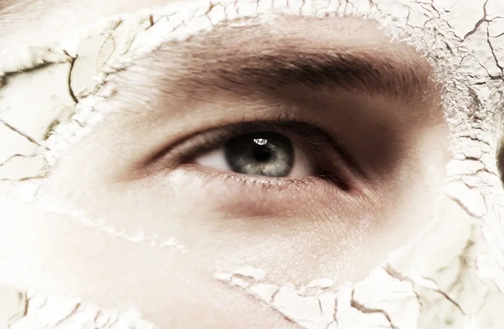 Close-up of a human eye peering through a hole in cracked, dry, white paint or plaster.