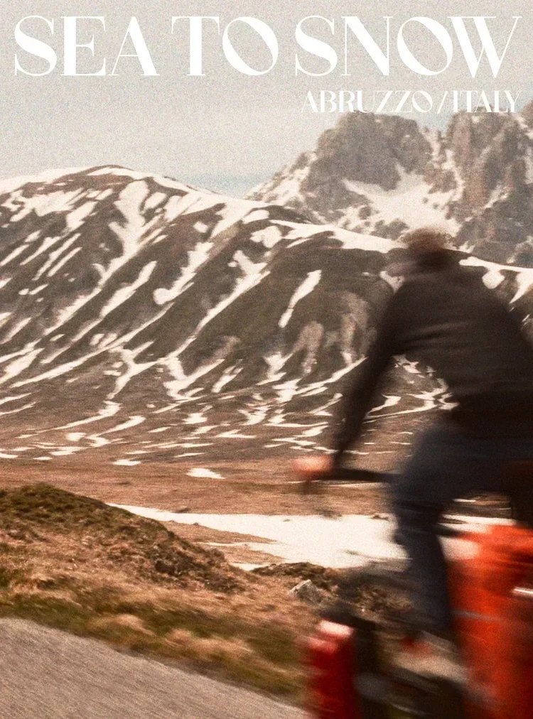 A person riding a bicycle on a mountain road with snow-capped peaks in the background, in Abruzzo, Italy.