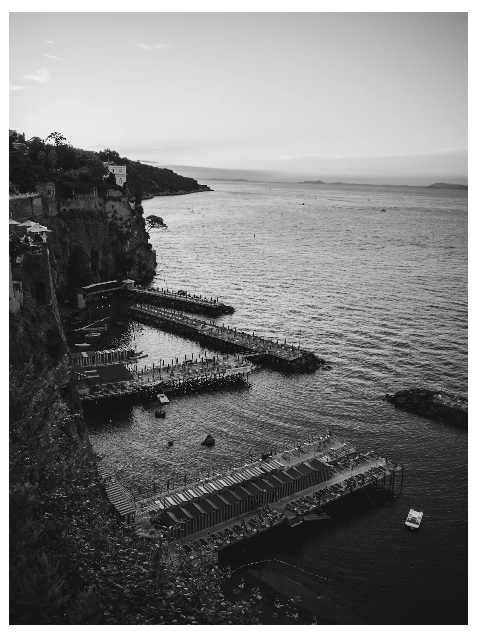 A black and white photo of a coastal area with a dock, boats, and buildings along the shoreline, with hills in the background.