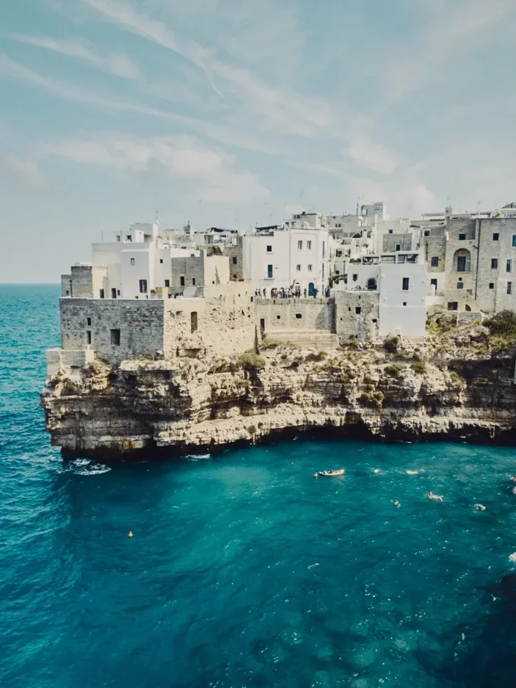 White buildings with flat roofs on a seaside cliff overlooking blue water, with a partly cloudy sky
