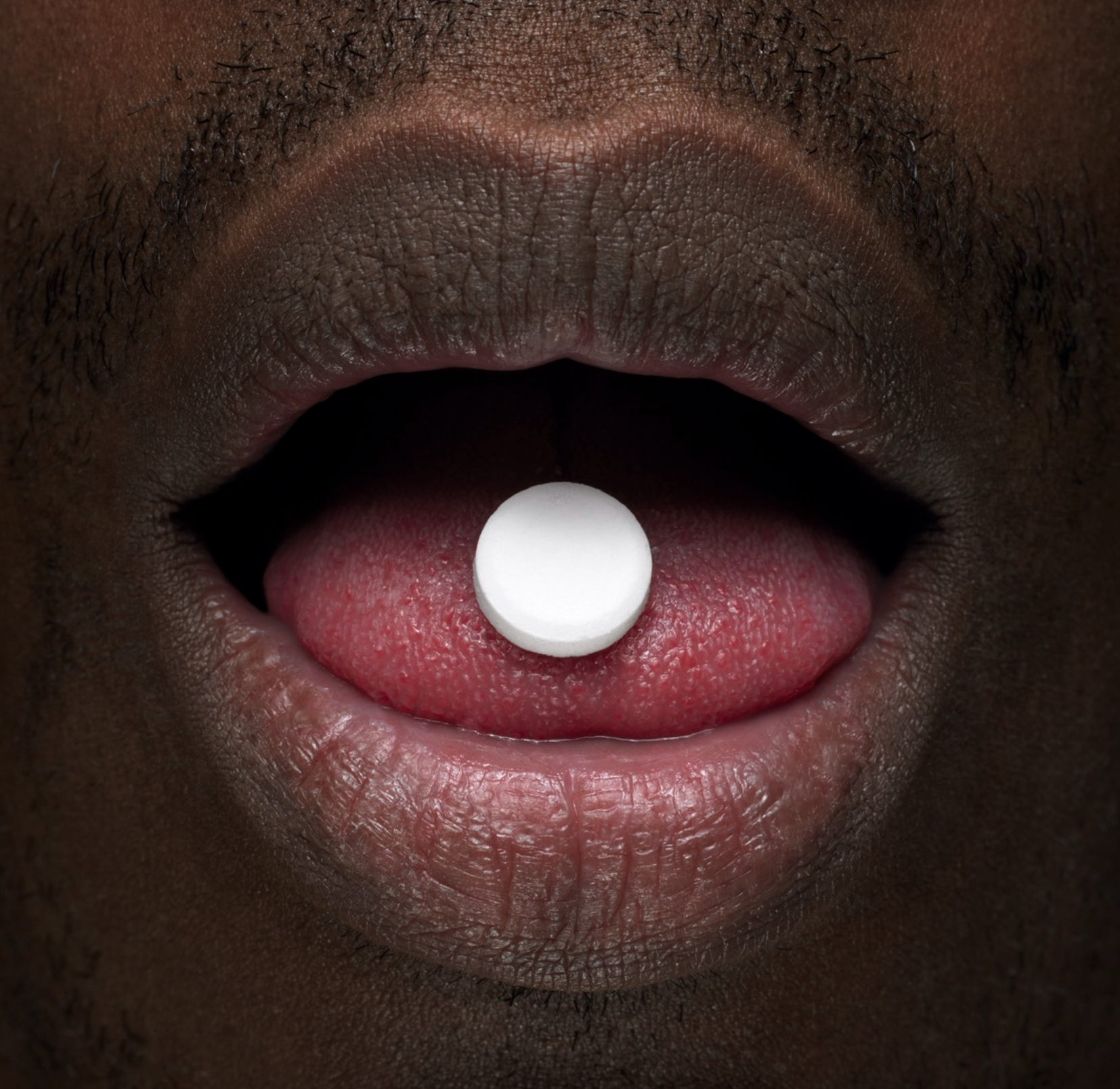 Close-up of a person's open mouth with a white pill resting on their pink tongue.