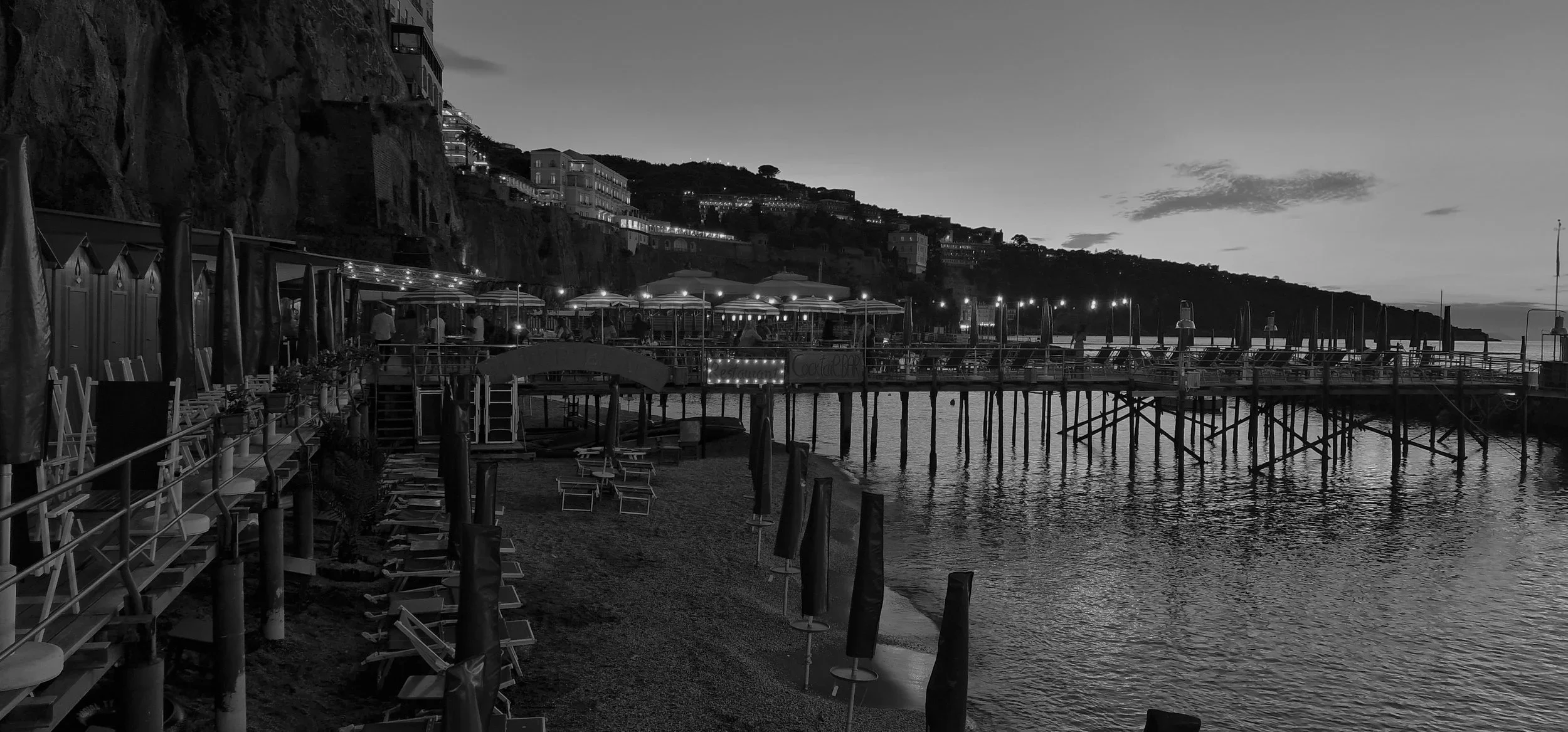 A black and white photo of a seaside pier with lounge chairs and umbrellas, buildings on a hillside in the background, and the water in the foreground.