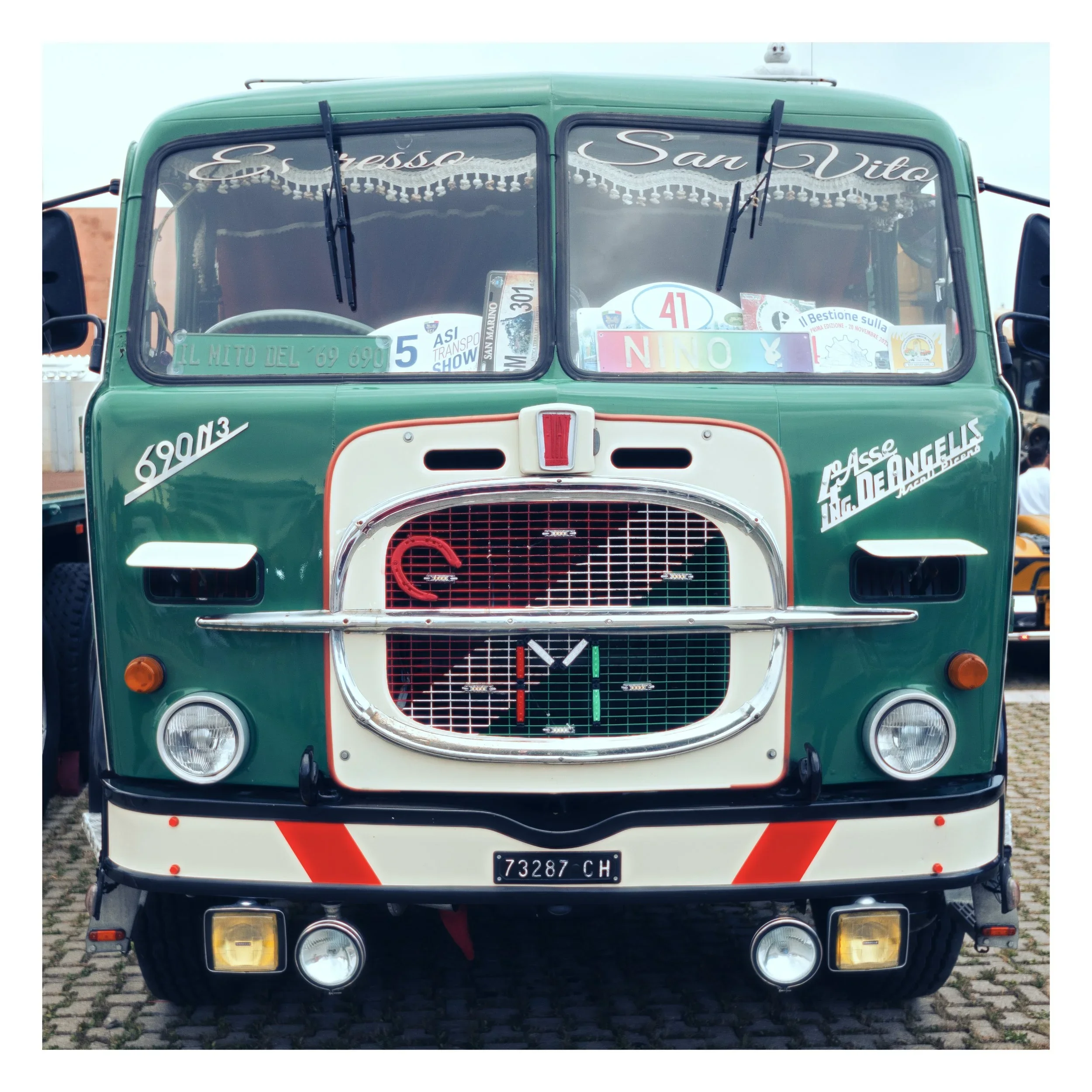 Front view of a vintage green bus with decorative window curtains and various stickers, parking on cobblestone pavement.