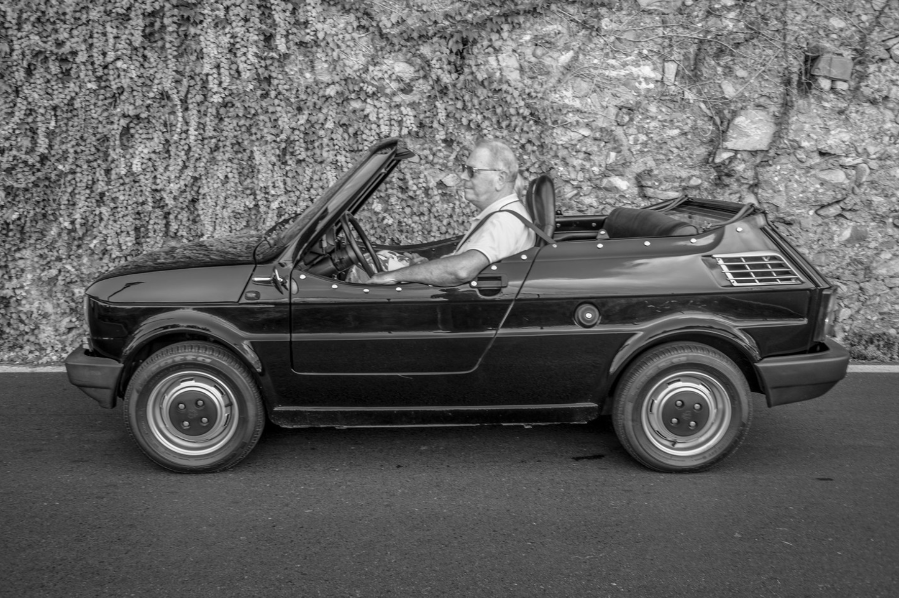 A man with sunglasses driving a small, vintage convertible car along a road with a rocky and leafy background.