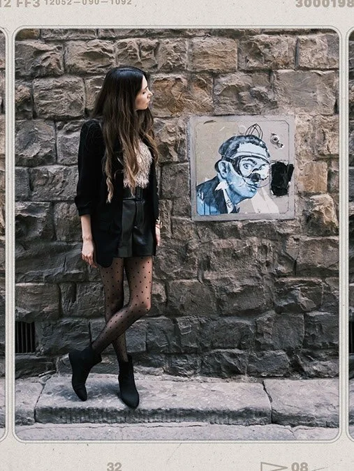 A woman with long hair standing against a stone wall, looking at a stencil graffiti of a person wearing glasses and headphones.