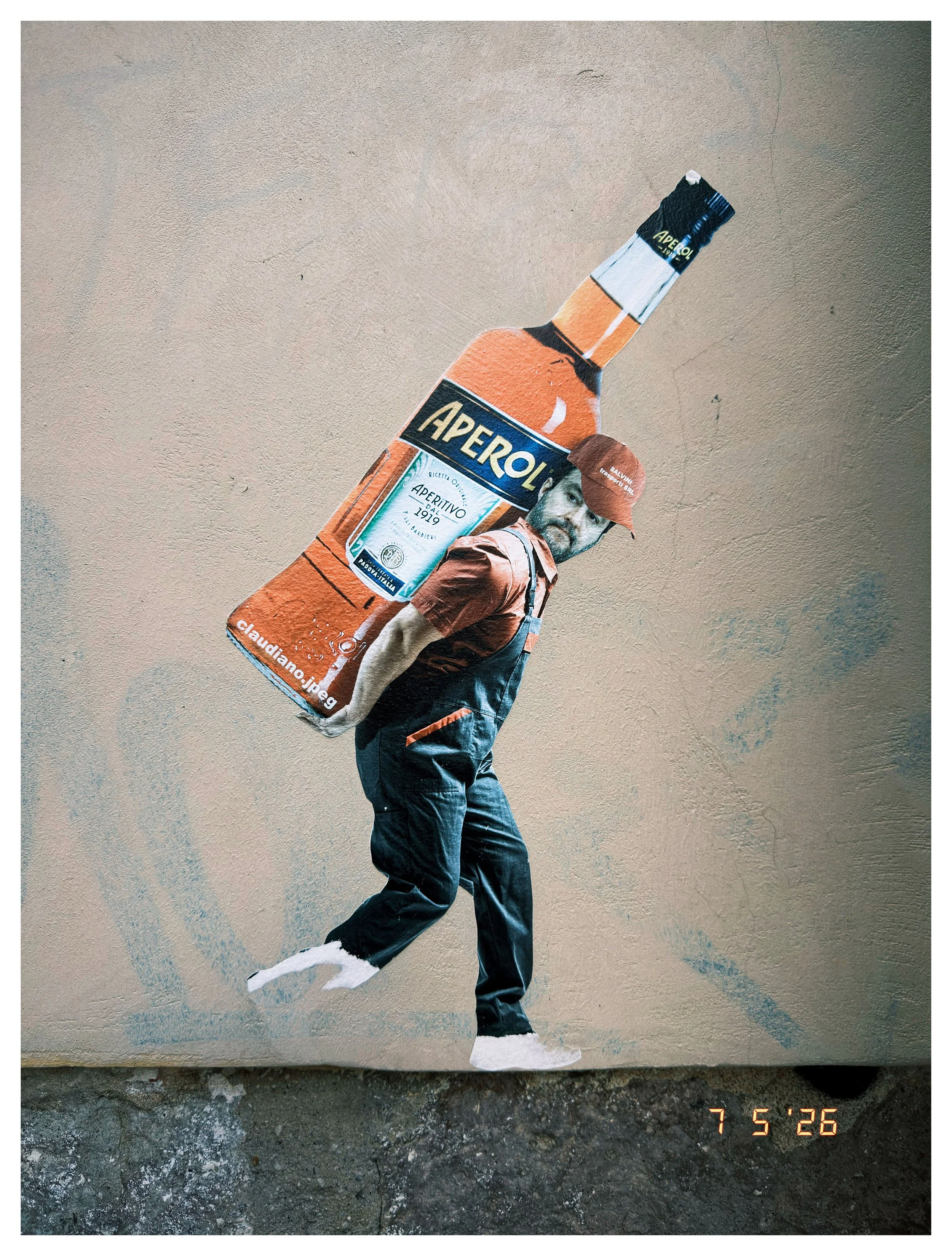 A street art mural of a person in casual clothing carrying a large bottle of Aperol soda, with the person's face partially hidden beneath the bottle, painted on a beige wall.