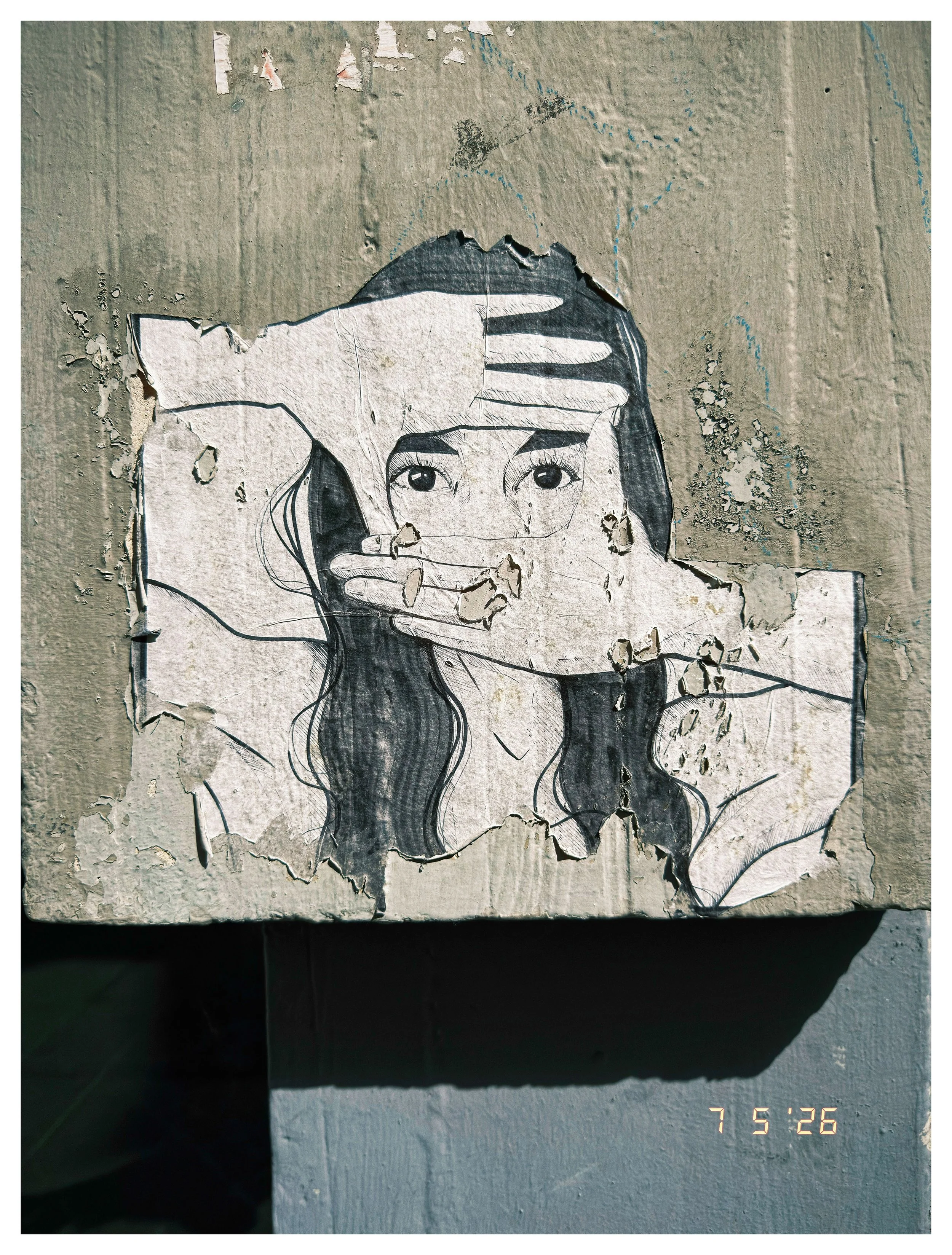 A black and white sticker of a woman's face with long hair, with her hand placed on her forehead, covering part of her eyes. The sticker is peeling and mounted on a textured wall with peeling paint and graffiti.