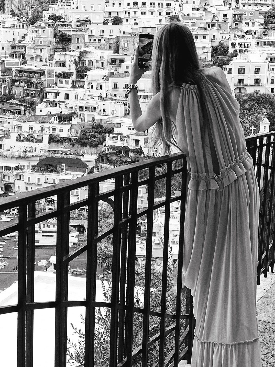 A woman in a flowing dress taking a photo with her smartphone on a balcony overlooking a hillside apartment area with numerous white buildings.
