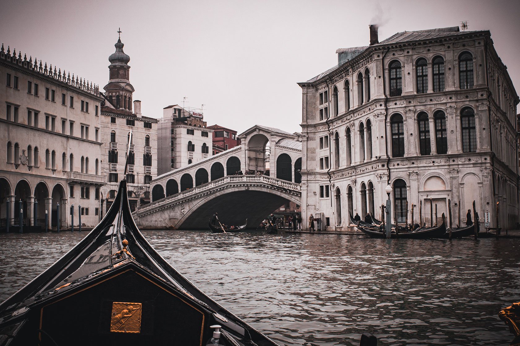 View from a gondola on a canal in Venice, Italy, showing historic buildings and the Rialto Bridge over the water.
