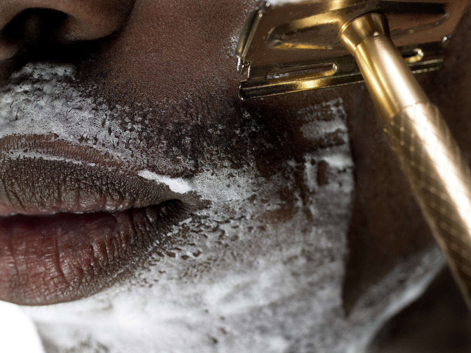 Close-up of a person's face with wrinkles, dry skin, and some white foamy soap. Part of a metallic grooming tool is visible near the face, suggesting a shaving or grooming context.